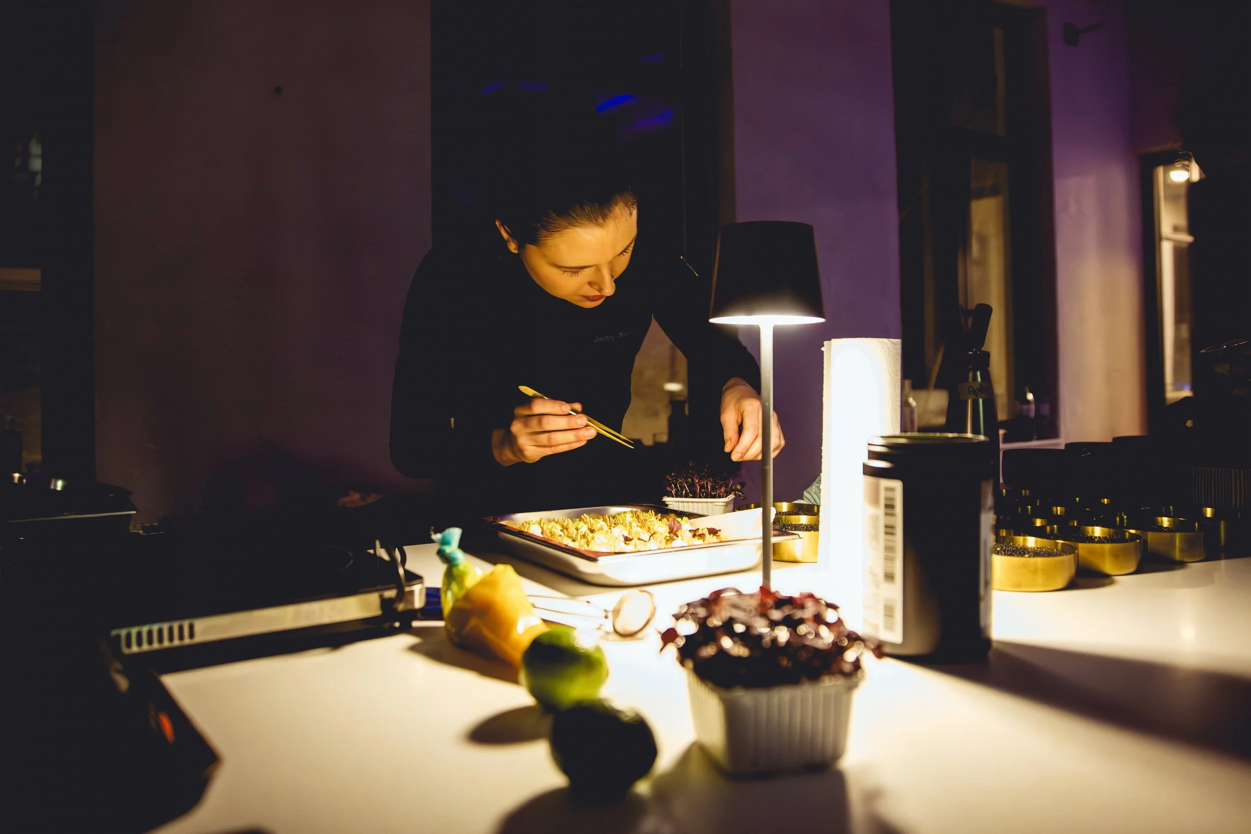 Jaimy Reisinger preparing sushi at a dimly lit counter with a black lamp and various ingredients.
