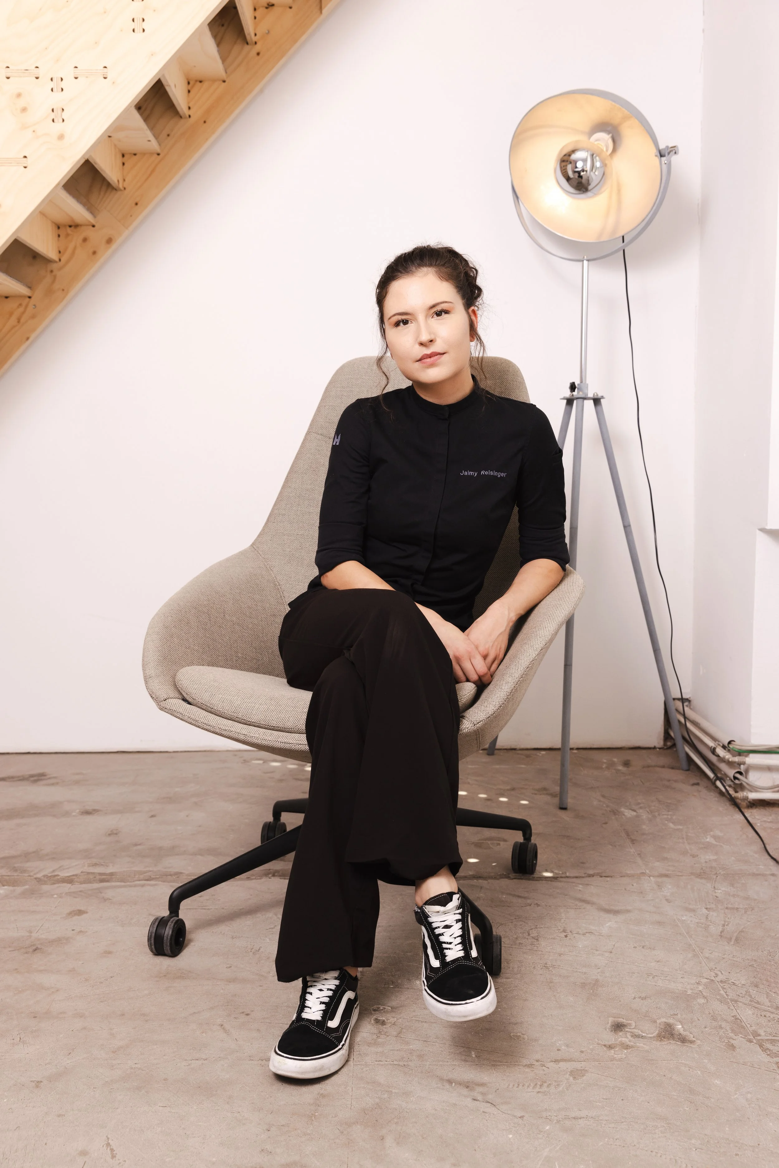 Jaimy Reisinger sitting in a beige office chair in a room with a wooden staircase and a large floor lamp behind her. She is wearing a black chef's coat, black pants, and black and white sneakers, looking at the camera with a neutral expression.
