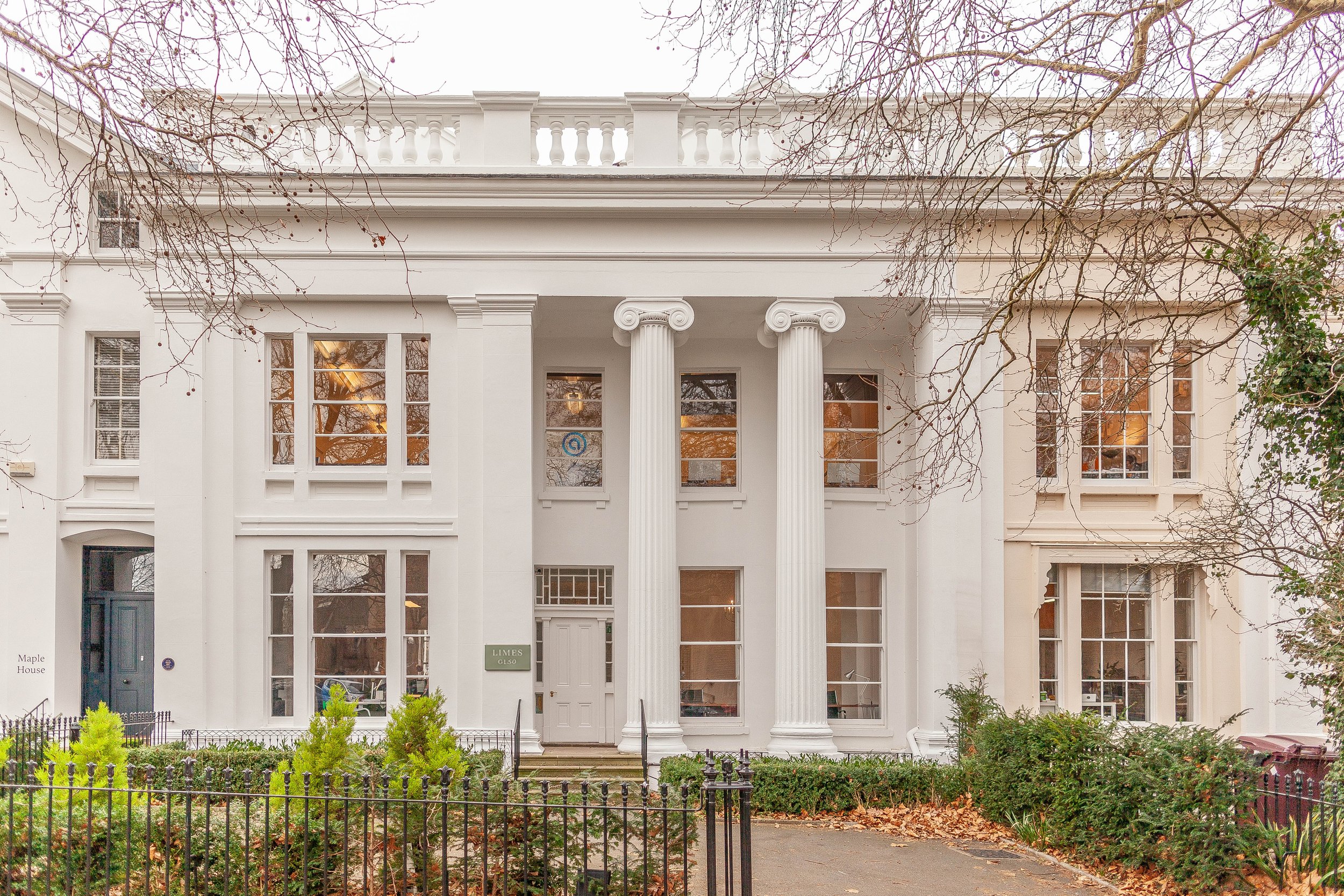 Front view of a white, two-story building with classical architecture including tall columns, large windows, and decorative trim, surrounded by a small garden and a black wrought iron fence.