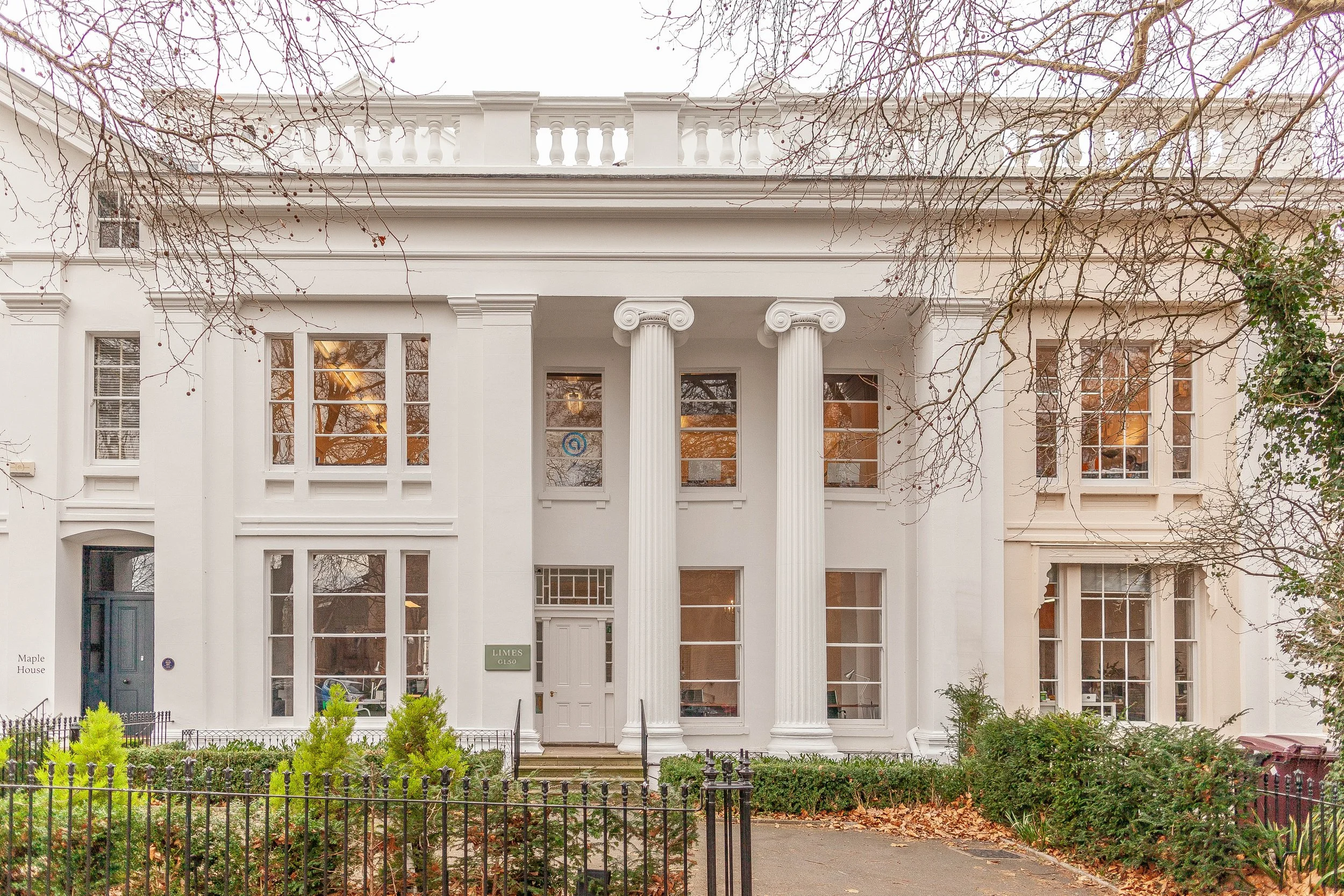 White neoclassical house with three prominent columns at the entrance, multiple windows, and decorative railing on the balcony. There are leafless trees with some red berries and a small garden with a black iron fence in the front.