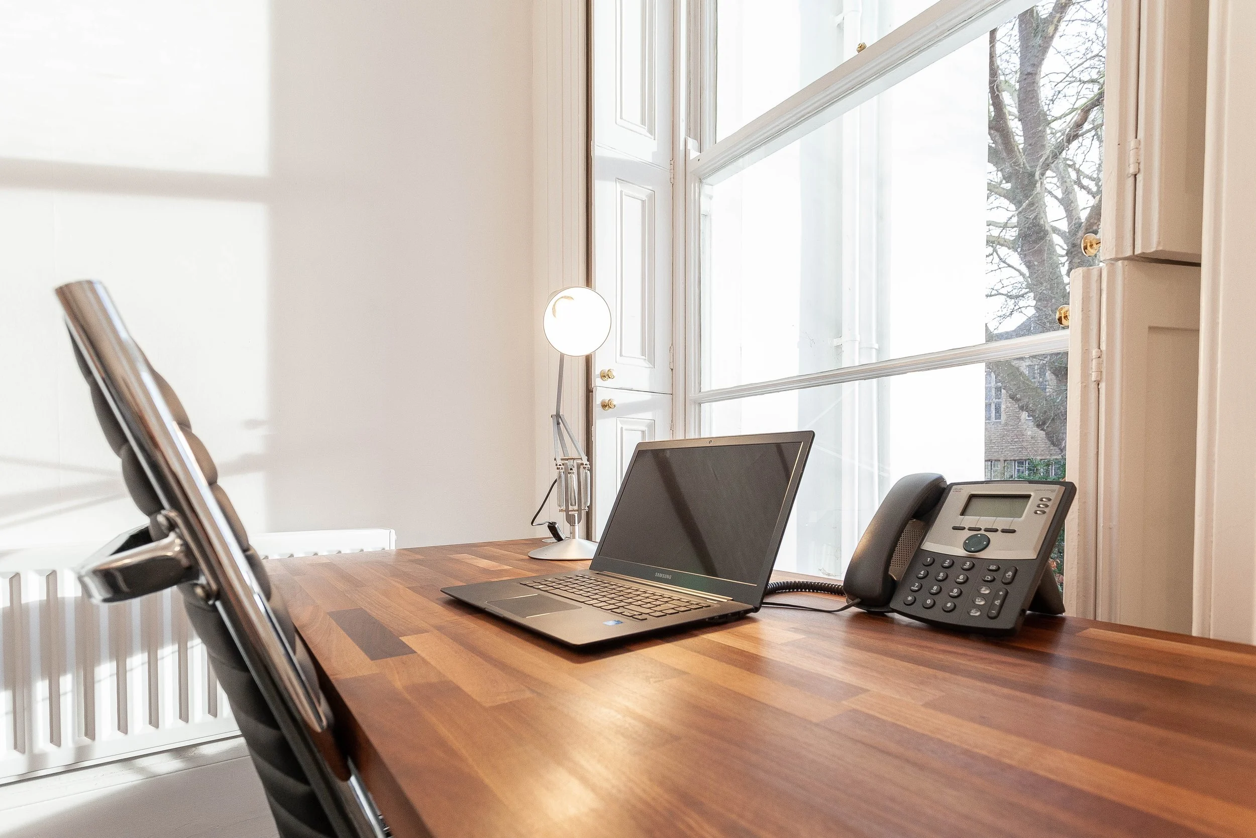 Office workspace with a wooden desk, a laptop, a desk phone, a modern reading lamp, and chairs, with large windows and a view of trees outside.