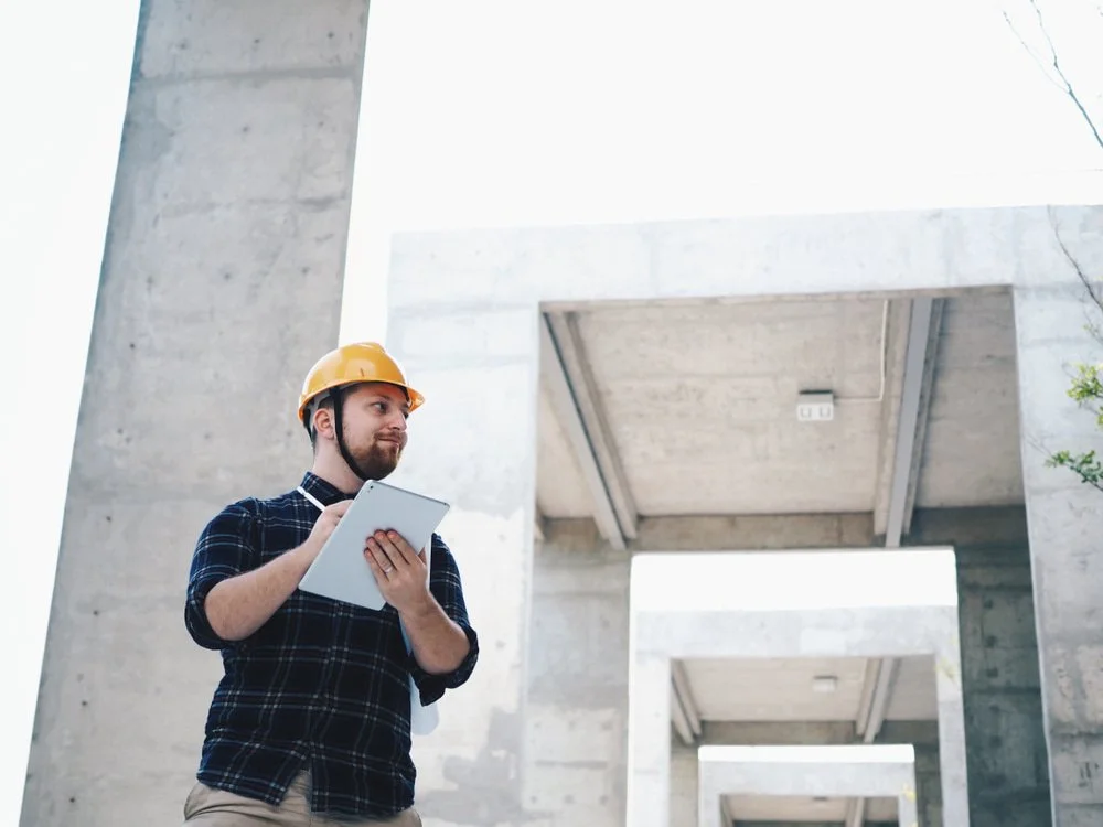 A man wearing a yellow construction helmet and plaid shirt holding a tablet, standing at a modern concrete building site.