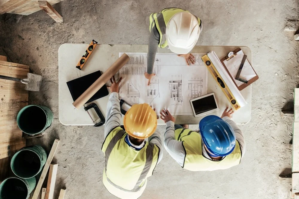 Three construction workers wearing safety helmets gathered around a table reviewing blueprints and construction plans at a building site.
