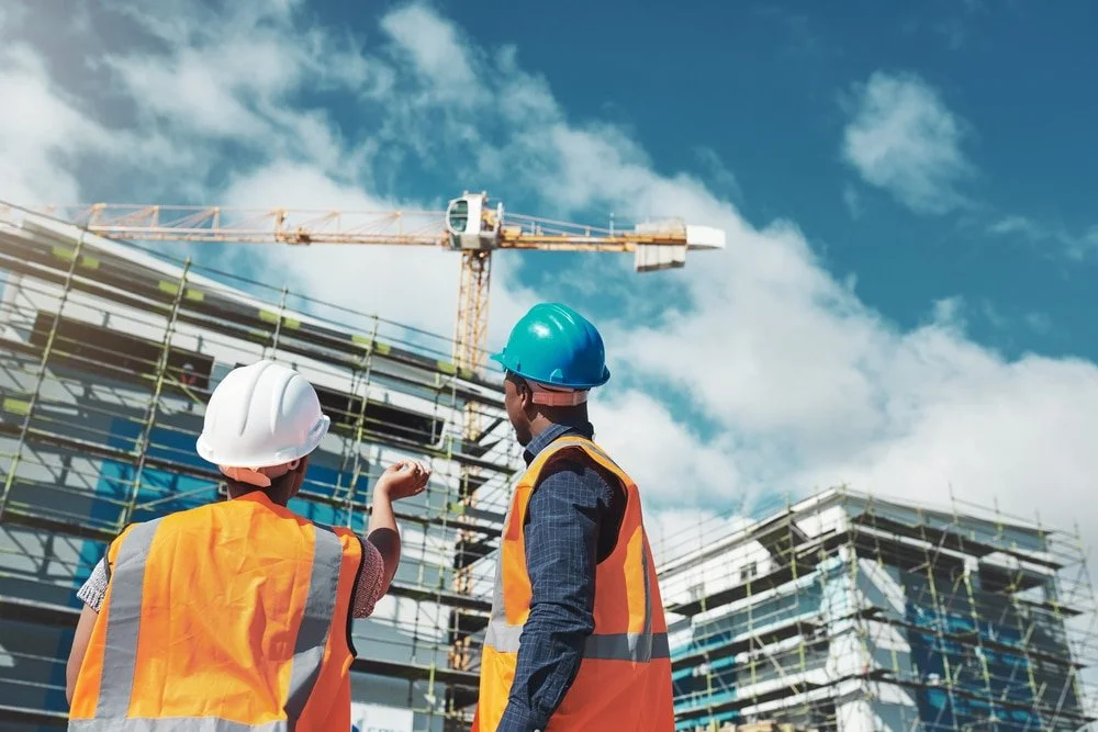 Two construction workers, wearing safety helmets and orange safety vests, standing on a construction site with scaffolding and a crane in the background, discussing progress.