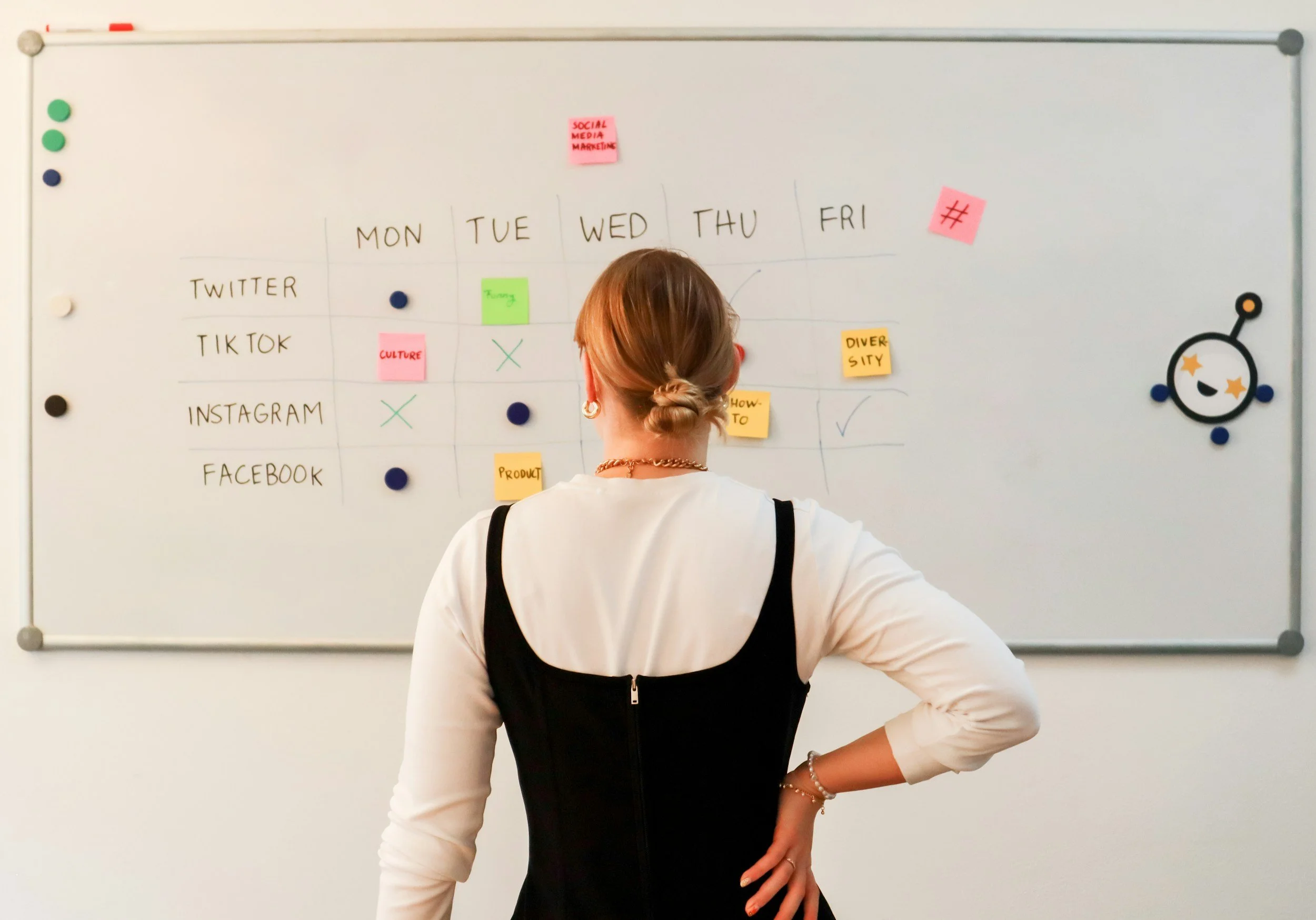 A woman with light brown hair in a braided bun is standing in front of a whiteboard with colorful sticky notes and handwritten text. The woman is wearing a black and white top with gold jewelry.