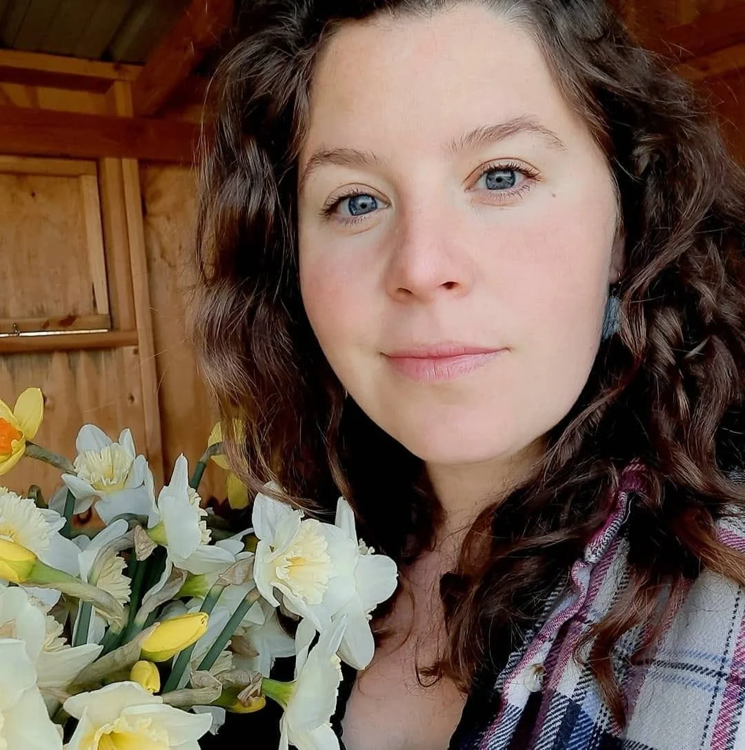 Close-up of a woman with curly brown hair and blue eyes, holding a bouquet of white and yellow daffodils, inside a wooden shed or barn.