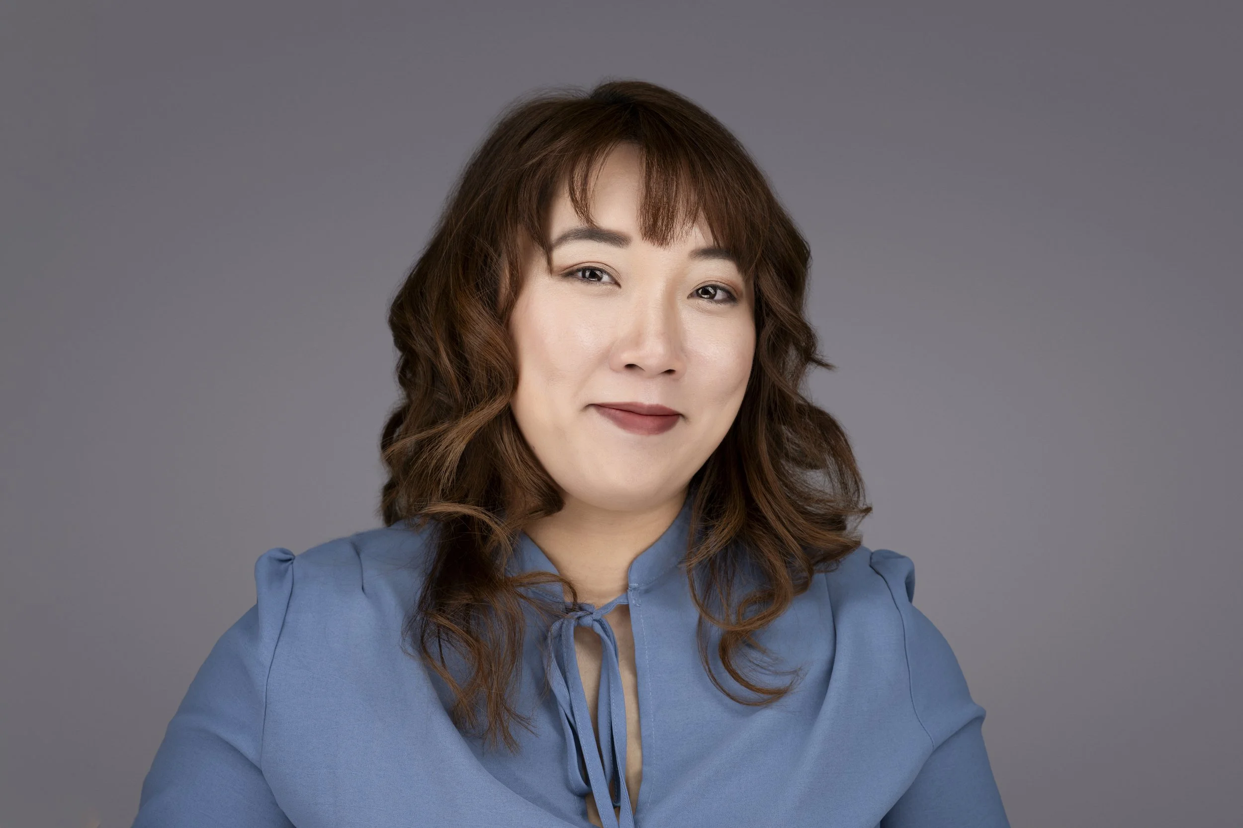 A woman with shoulder-length wavy brown hair, wearing a blue blouse, looking at the camera with a subtle smile against a plain gray background.