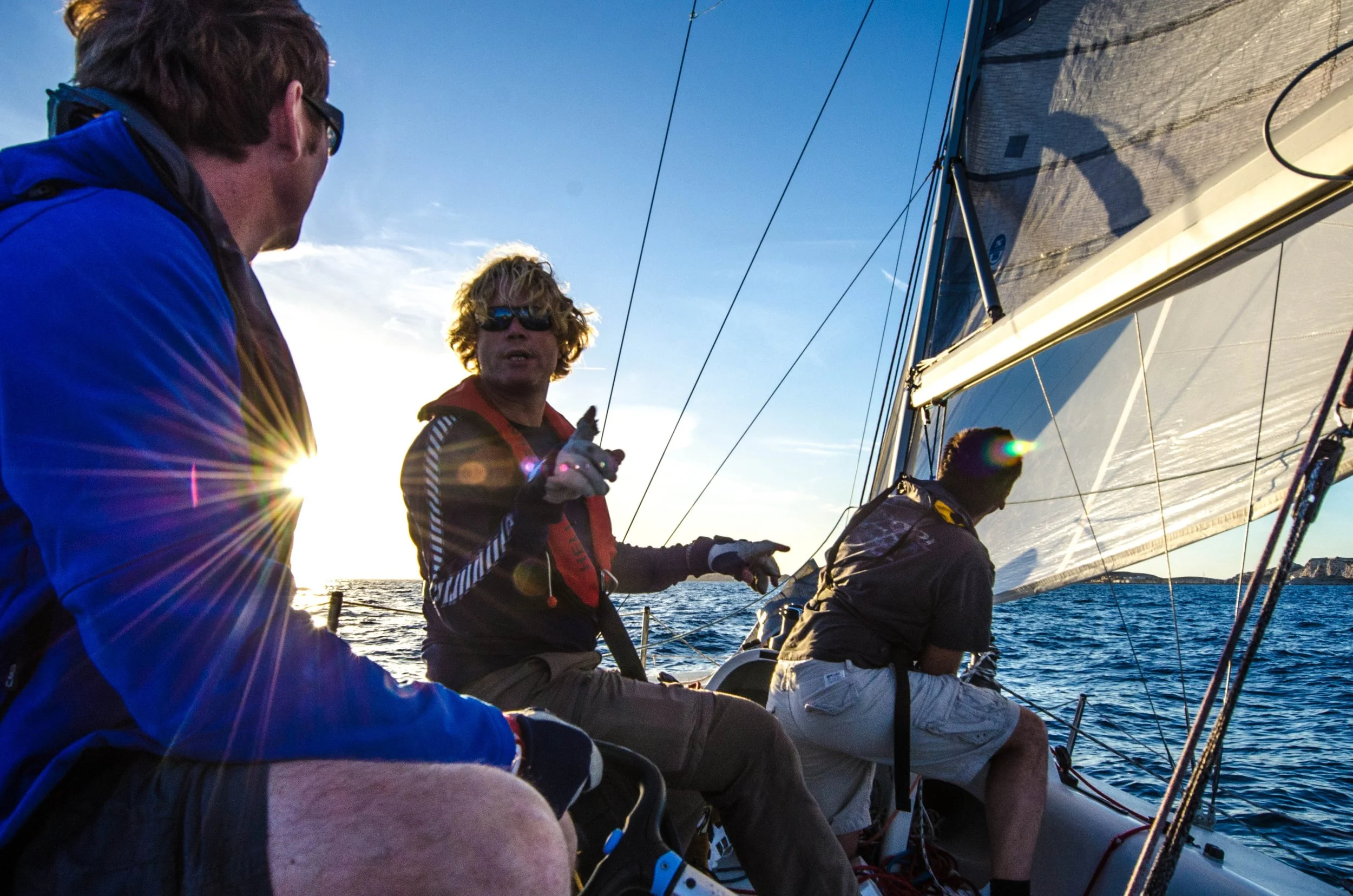 People sailing on a boat during sunset, with a man in a blue jacket and others tending to the sail, with the sun and water in the background.