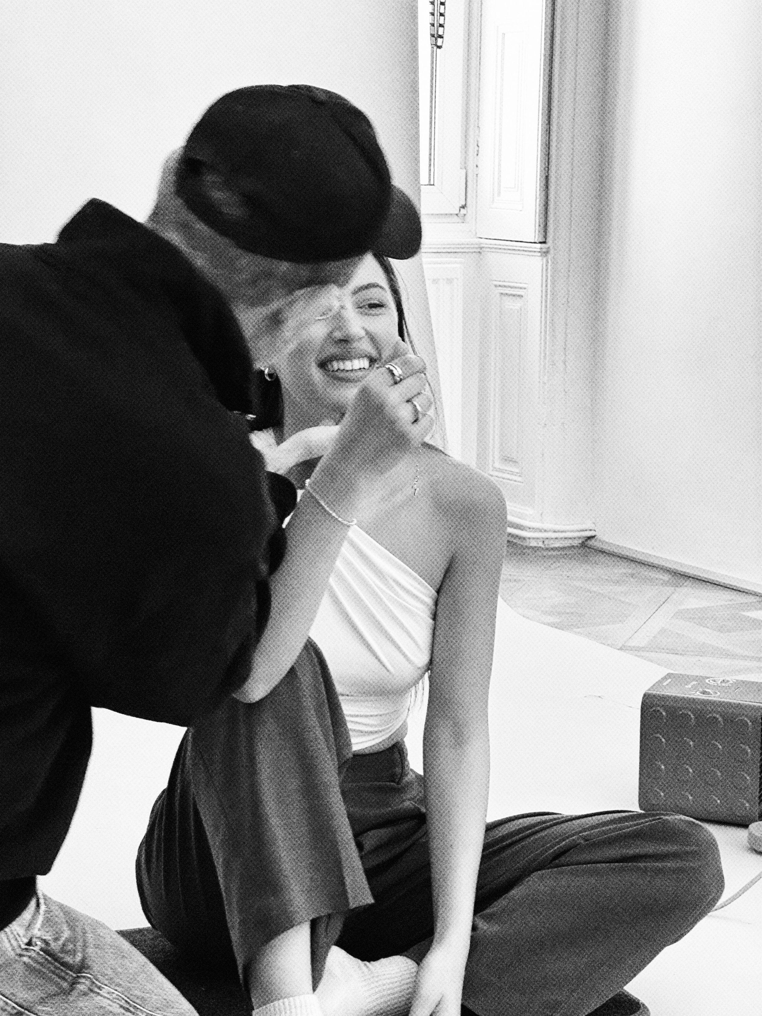 A woman sitting on the floor while a person applies makeup to her face. The woman is smiling and wearing a strapless top and dark pants. The person applying makeup is wearing a cap, glasses, and a jacket.