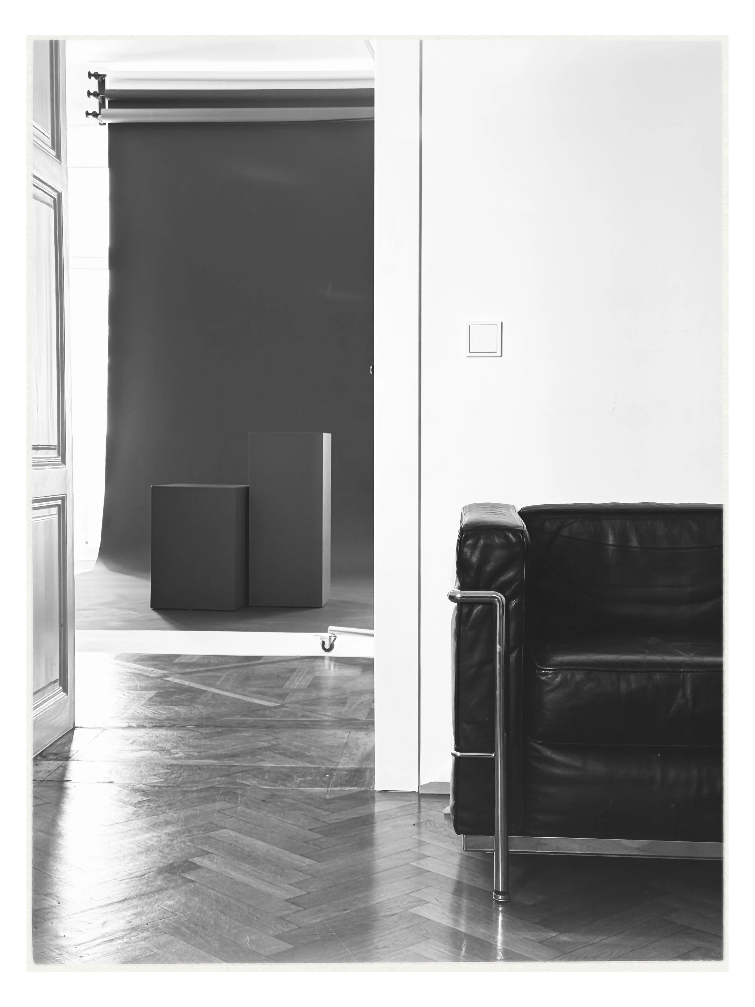 A black leather couch in a room with wooden parquet flooring, a white wall with a switch, and an open doorway leading to a room with a dark backdrop and two rectangular pedestals.