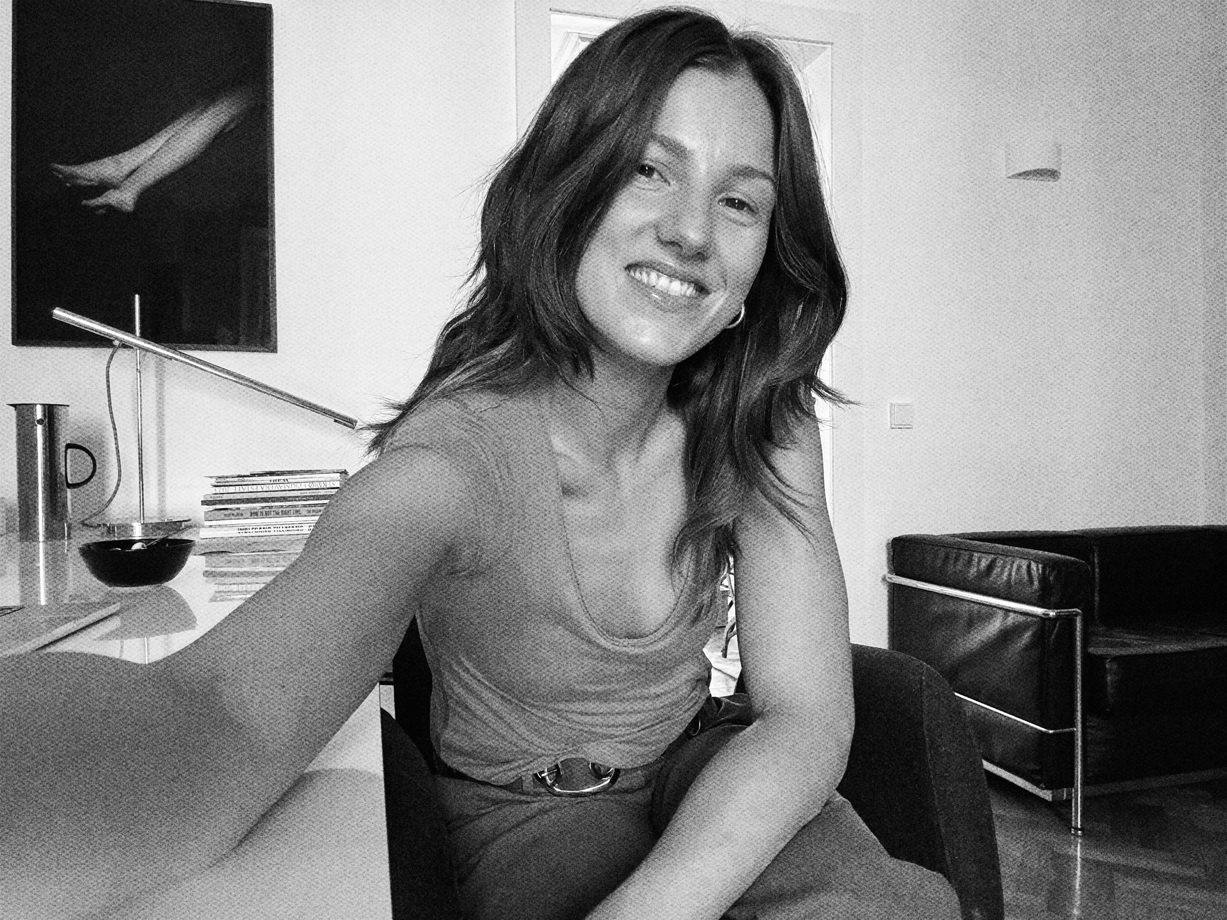 A woman with shoulder-length dark hair taking a selfie in a modern living room, smiling at the camera.