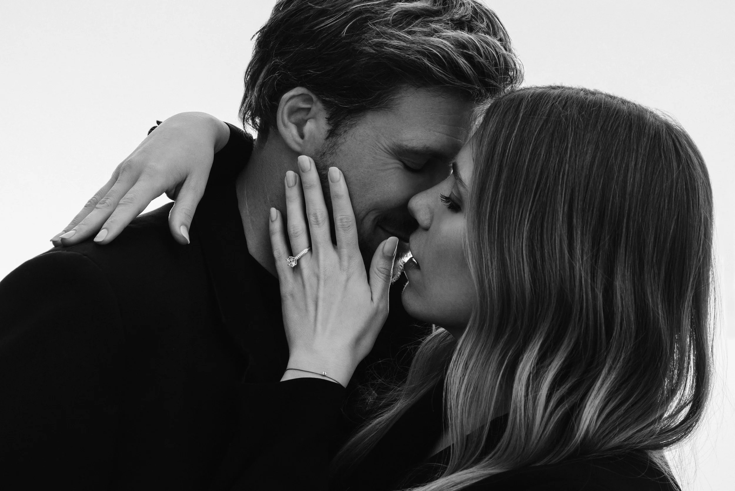 A black and white close-up photo of a couple about to kiss. The woman has her hand on the man's face, showing an engagement ring, and the man is leaning in with his eyes closed.