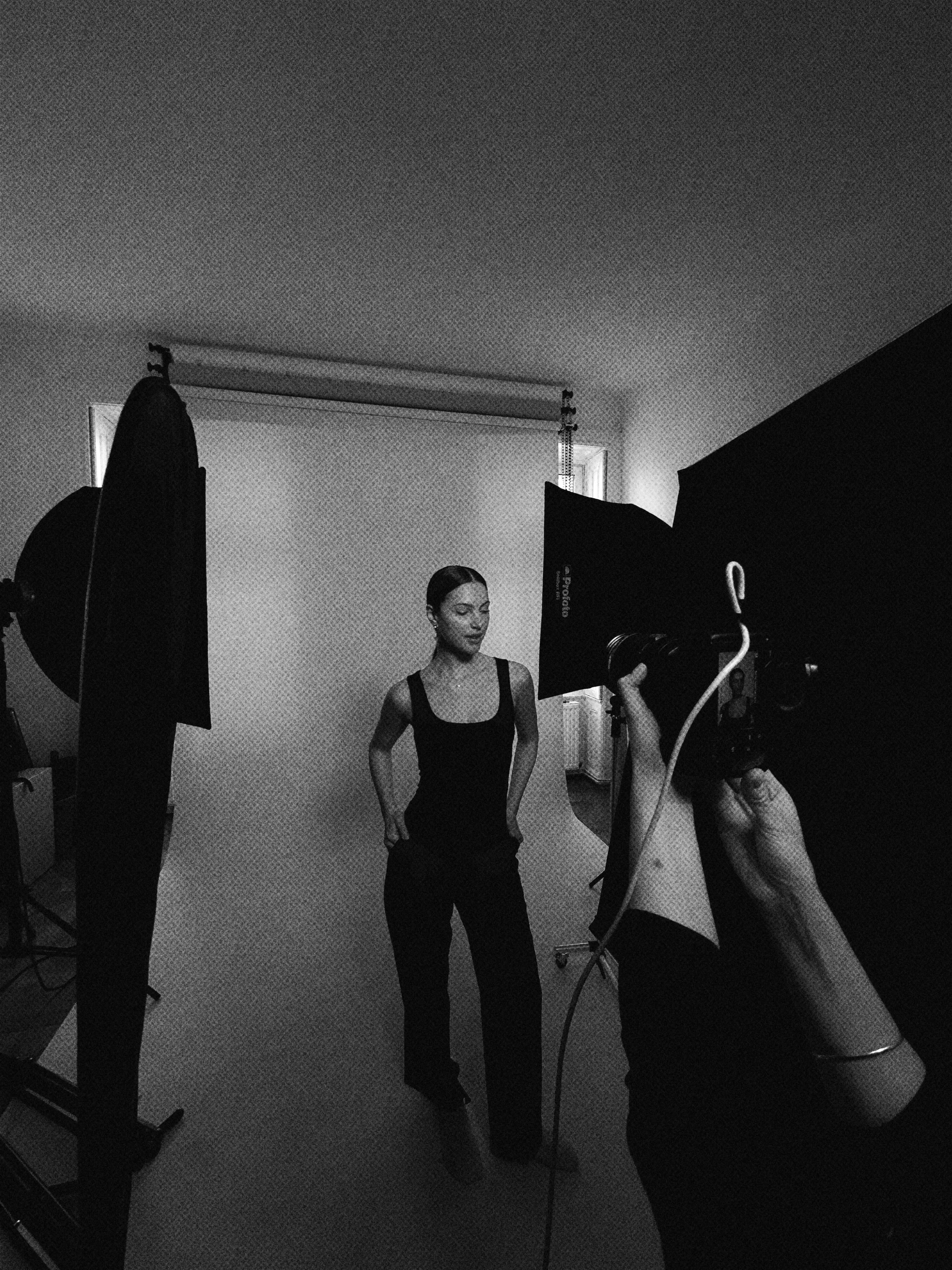 A woman in black clothing stands in front of a photography backdrop during a photo shoot, with photography equipment and lighting setup visible.