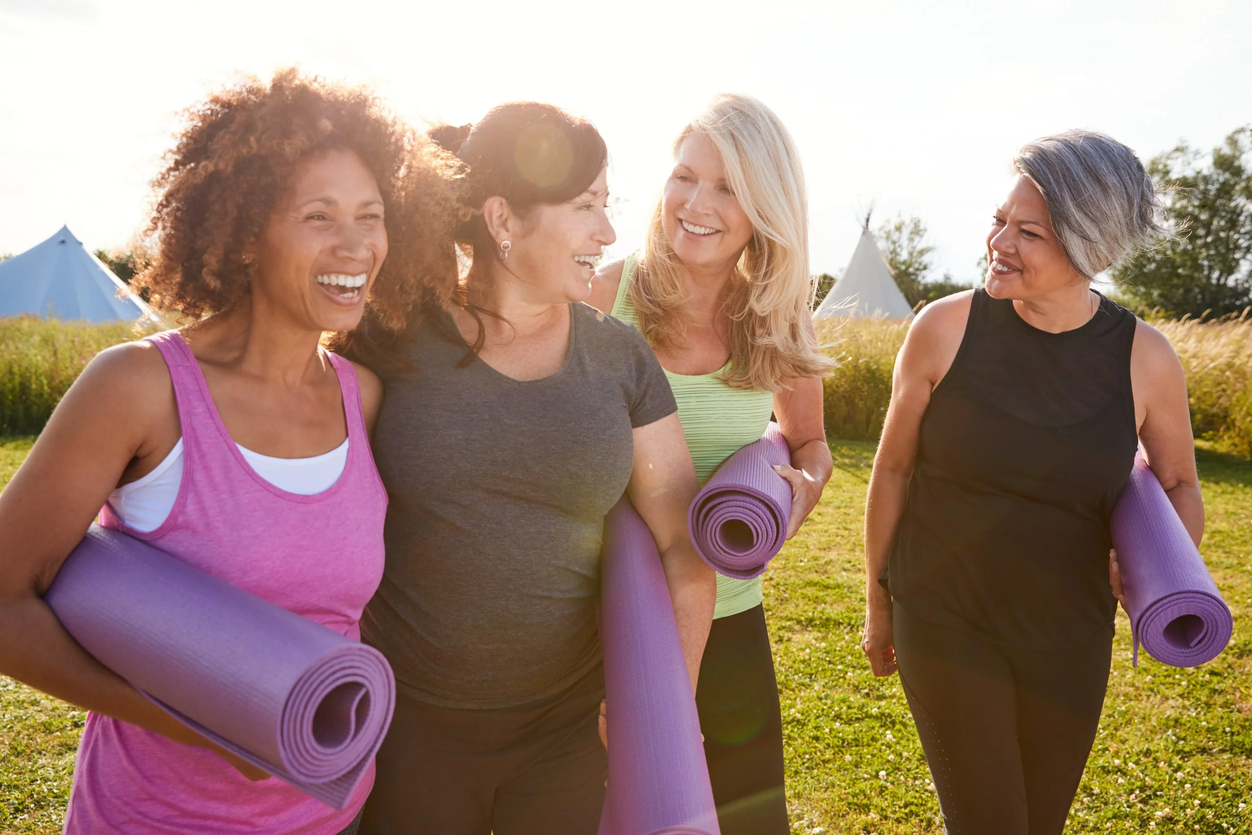 Four women smiling and carrying purple yoga mats outdoors during sunset.