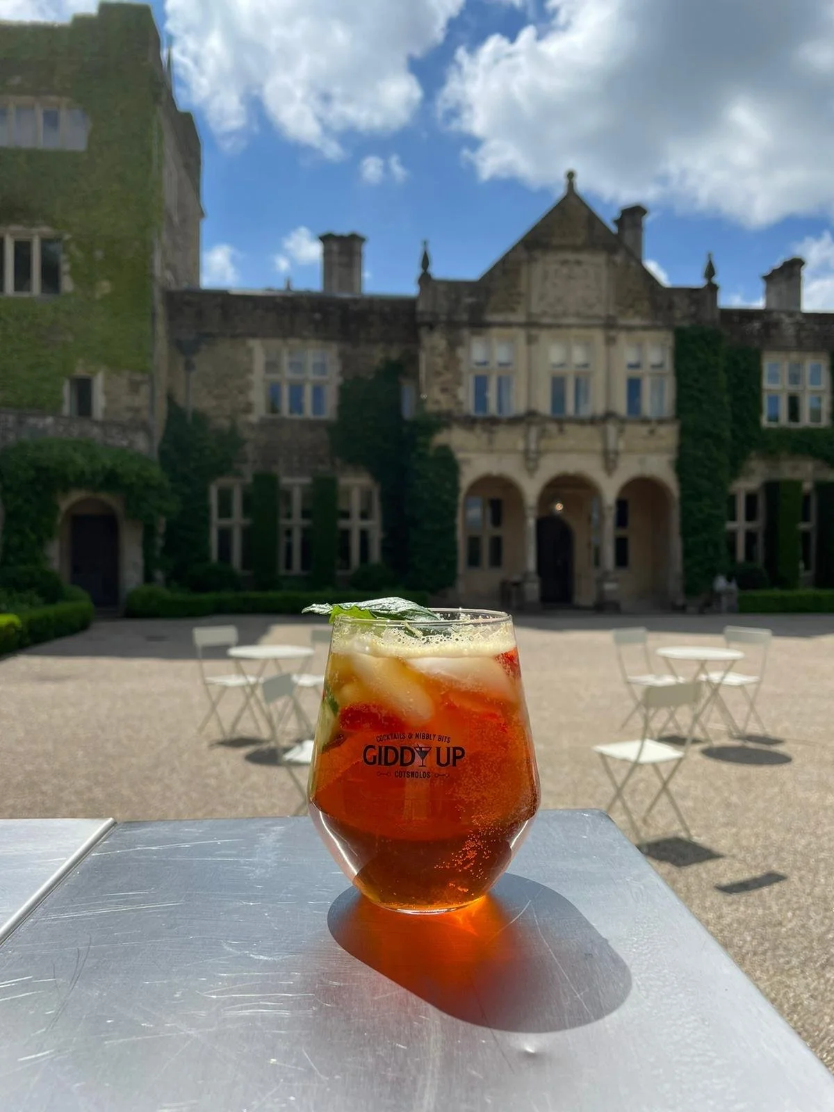 A glass of iced cocktail on a table outside with a historic stone building and a blue sky with clouds in the background.