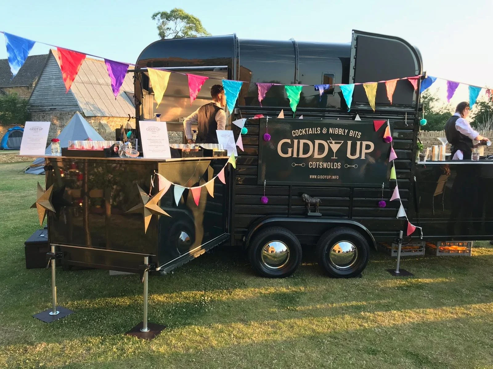 A black mobile bar called 'Giddy Up' serving cocktails at an outdoor event, decorated with colorful bunting and star-shaped ornaments, with tents and rustic buildings in the background.