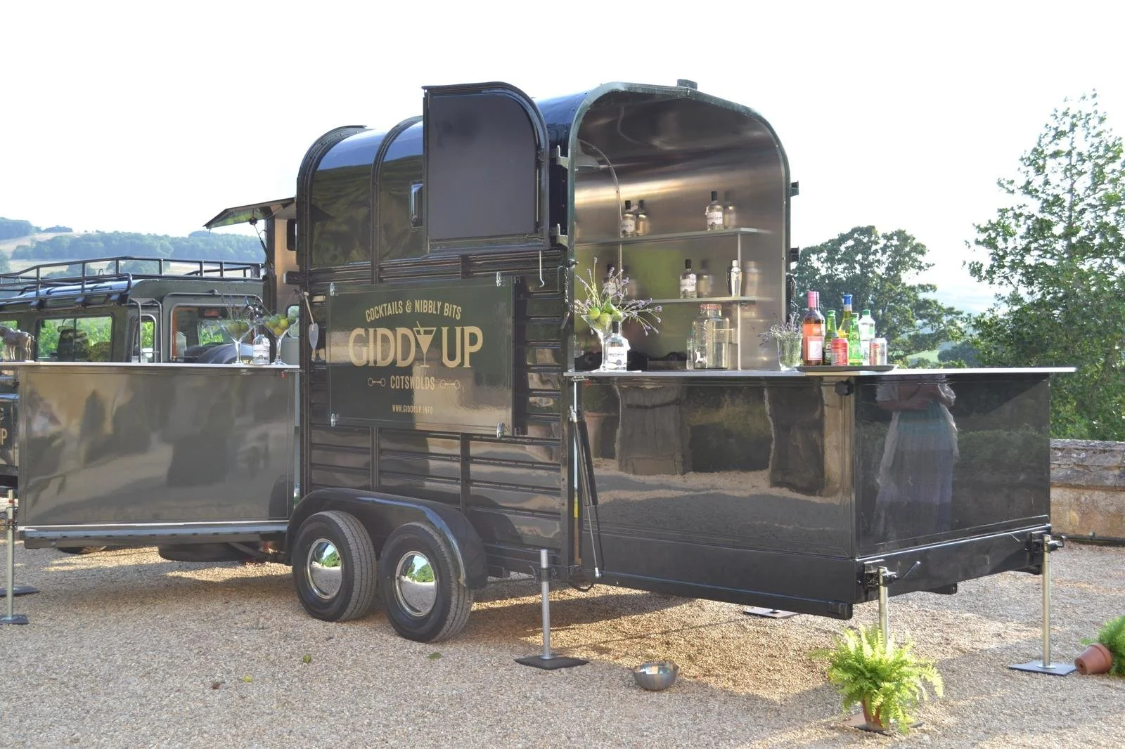 A black mobile cocktail bar trailer with the label "Giddy Up" offering drinks and garnishes outdoors on a gravel surface, with trees and hills in the background.