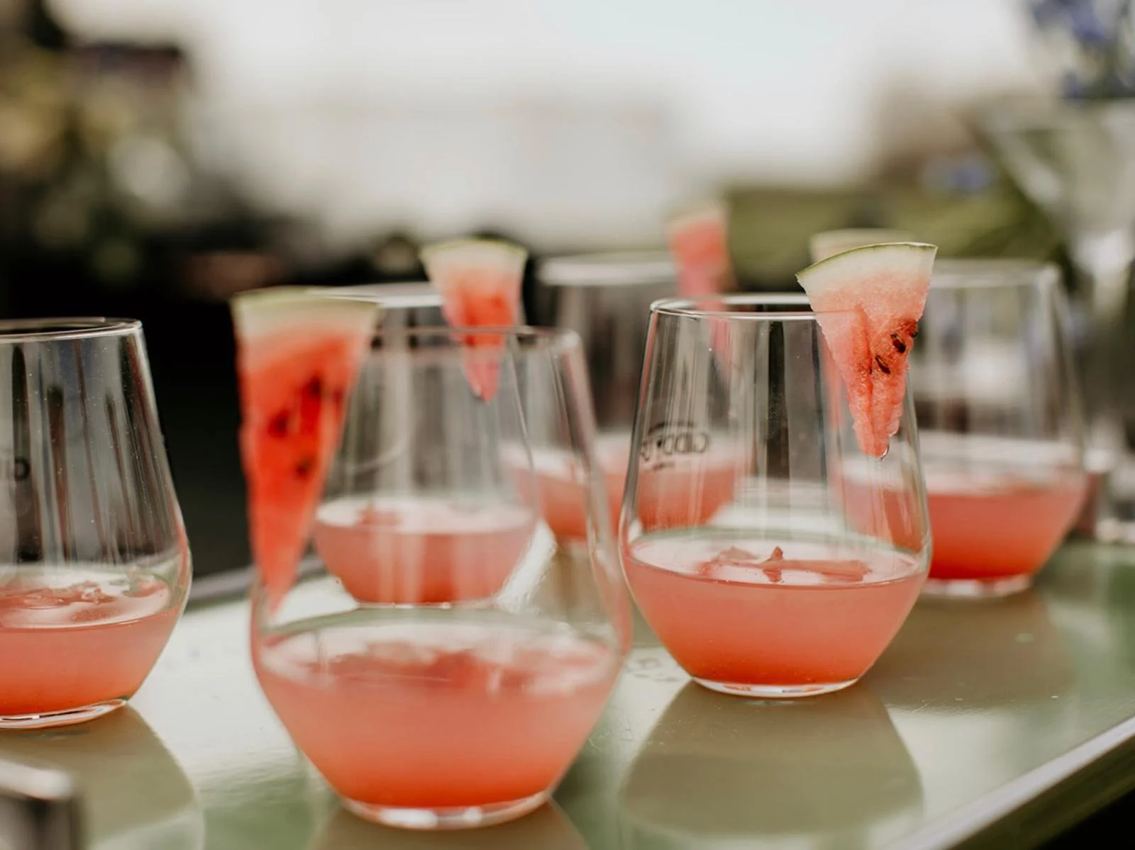 Several glasses with pink watermelon beverages, each garnished with a small wedge of watermelon, placed on a white surface.