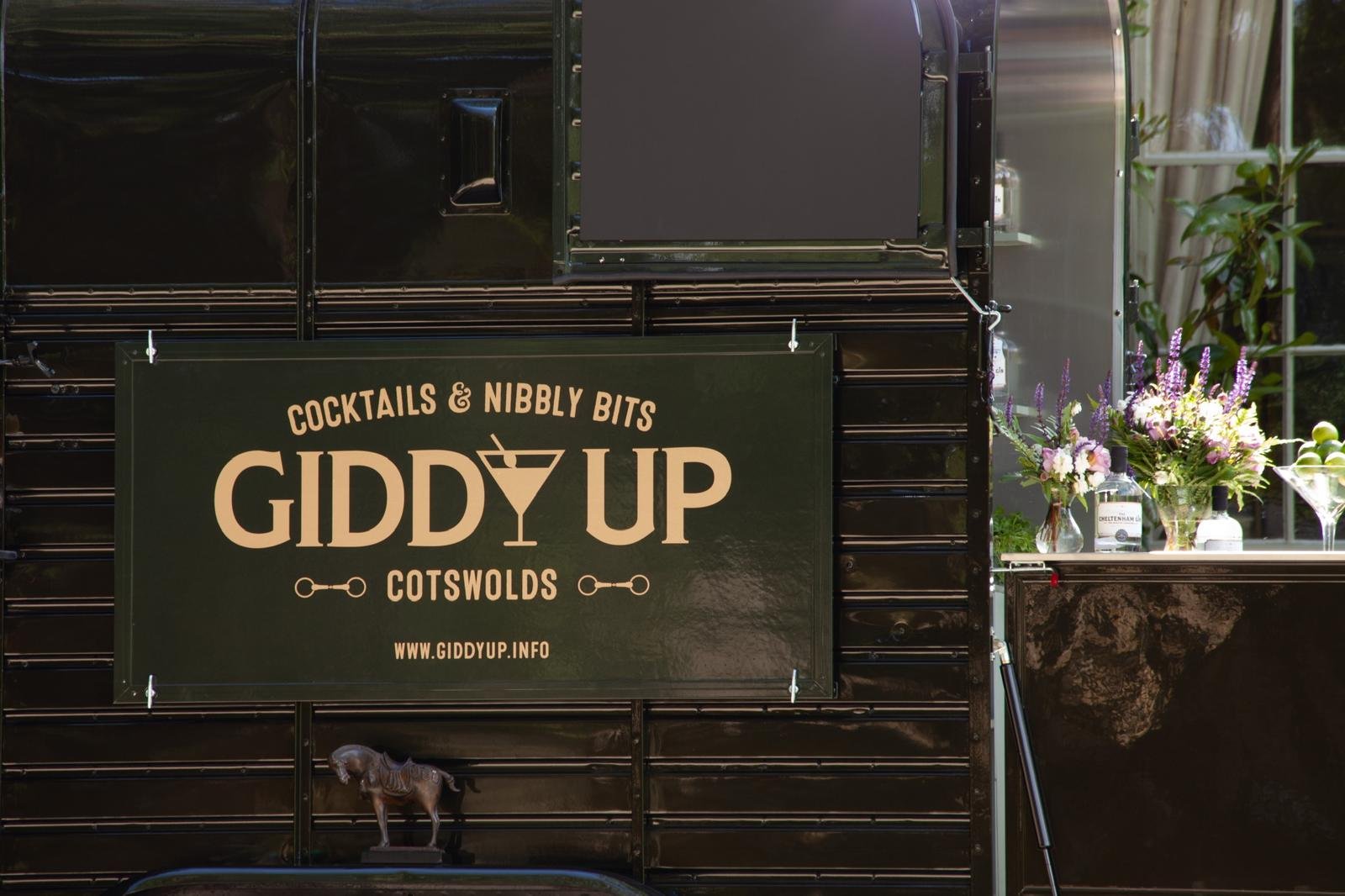 Black food truck with a sign that reads 'Cocktails & Nibbly Bits GIDDY UP COTSWOLDS' and a website link. The truck has a small horse figurine hanging at the bottom of the sign, purple and white flowers in vases, and bottles on a counter.