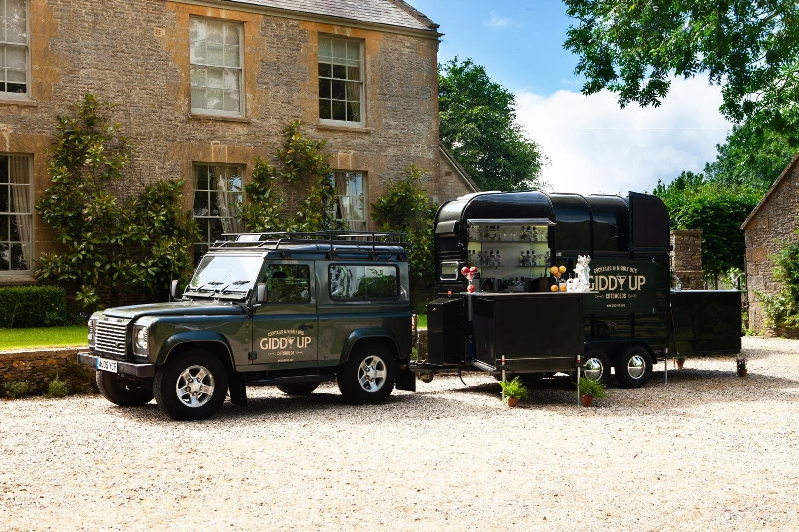 A black food truck shaped like a vintage Land Rover with the sign "Giddy Up" parked on a gravel driveway in front of an old stone building with large windows and greenery.