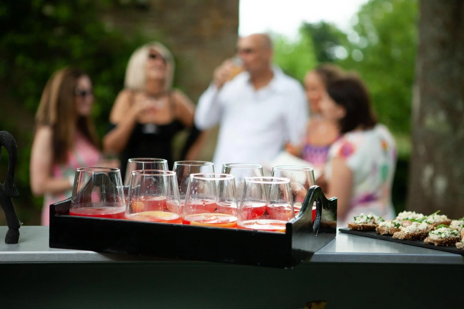 Tray of glasses filled with pink drinks on a table with blurred people in the background enjoying a social gathering outdoors.