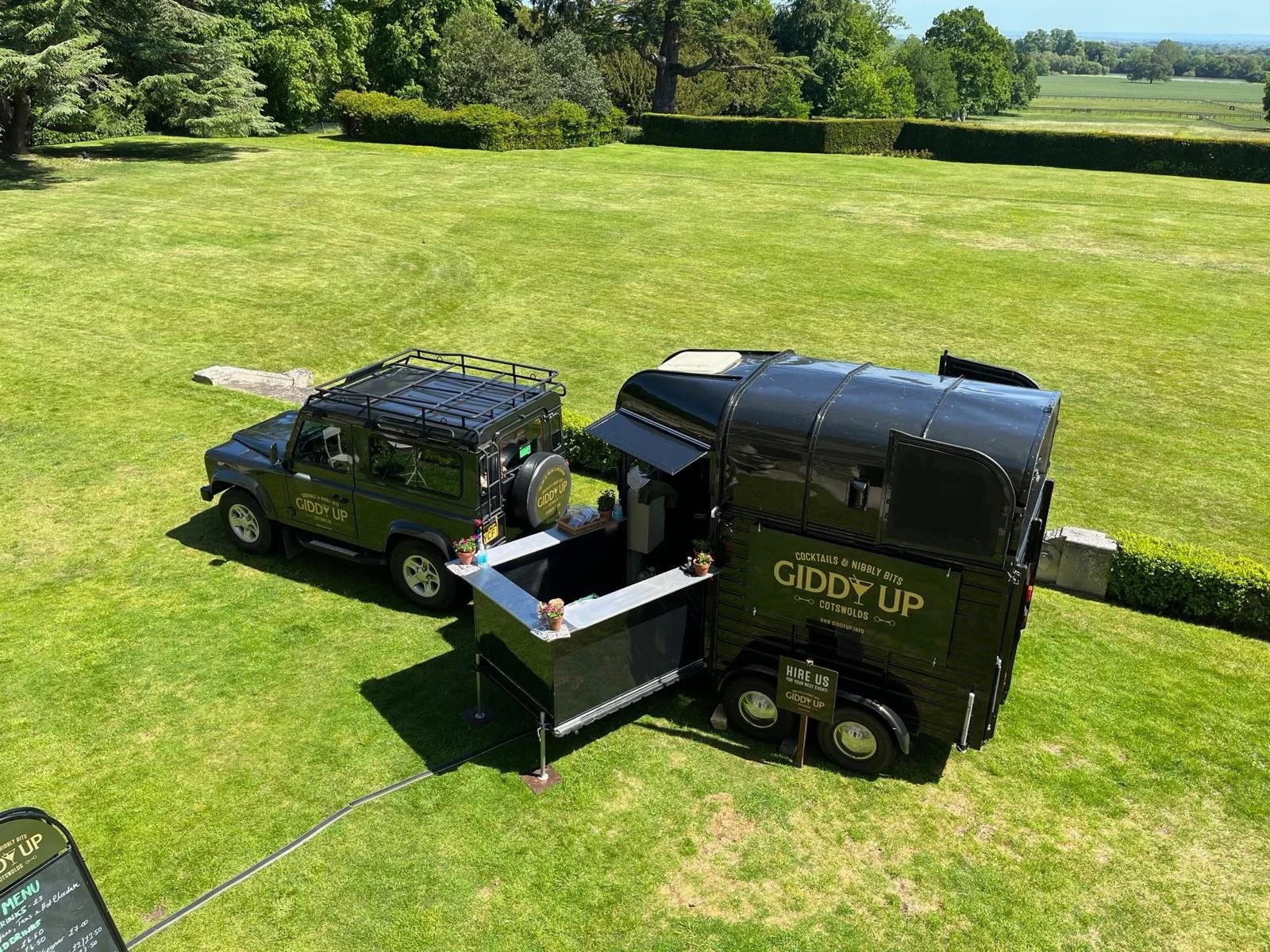 Black food truck with gold lettering, parked on a grassy field, selling cocktails and nibbly bites, with a smaller vehicle attached and a menu sign nearby.