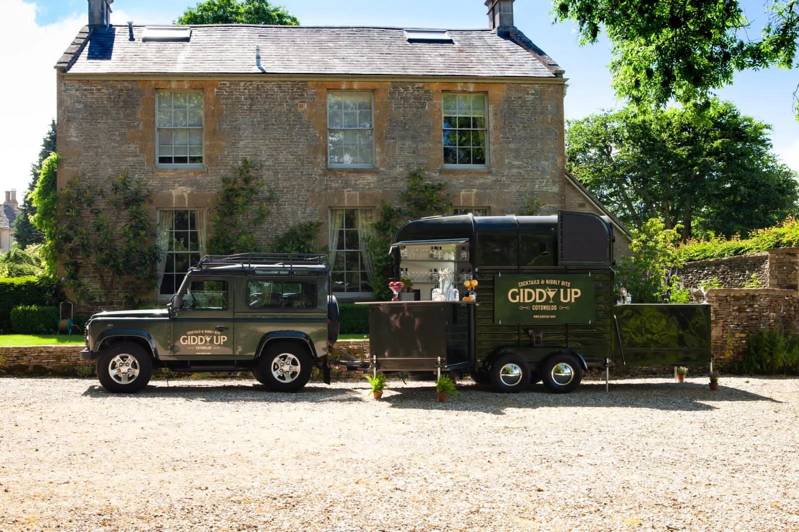 A black mobile bar trailer with the label 'Giddy Up Cocktails & Nibbly Bits' parked in front of a large brick house with trees and greenery.