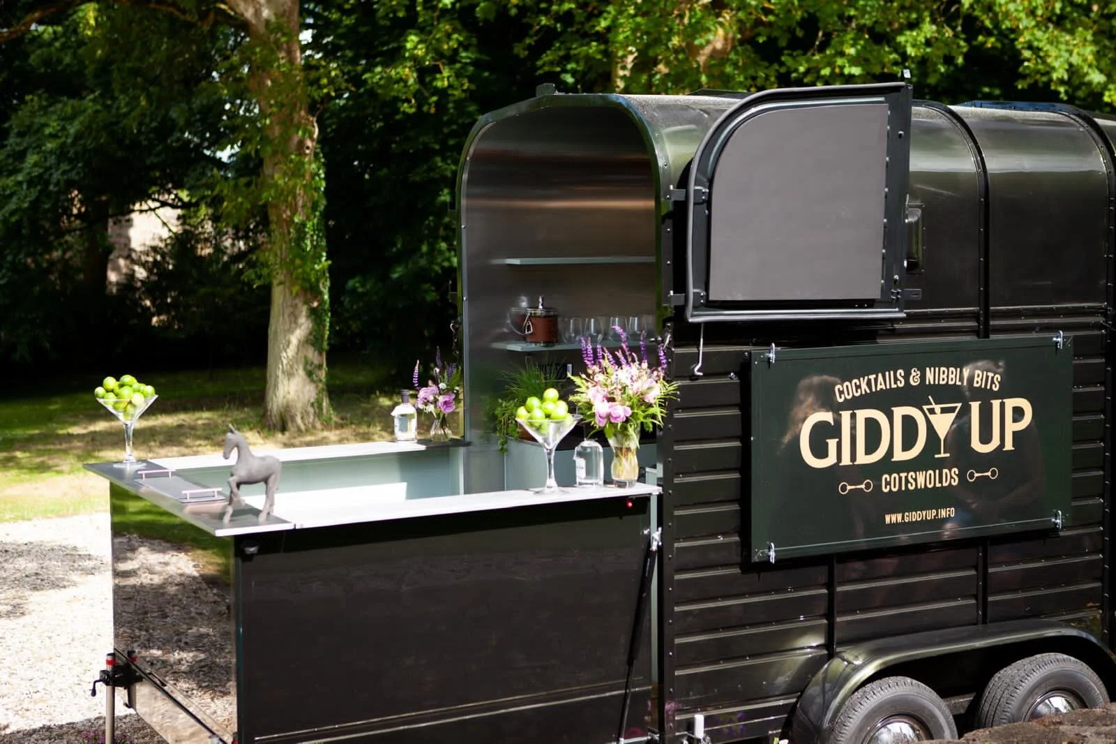 A black mobile bar cart with a sign that reads 'Giddy Up Cotswolds' with flowers and a green apple display, set outdoors with trees in the background.