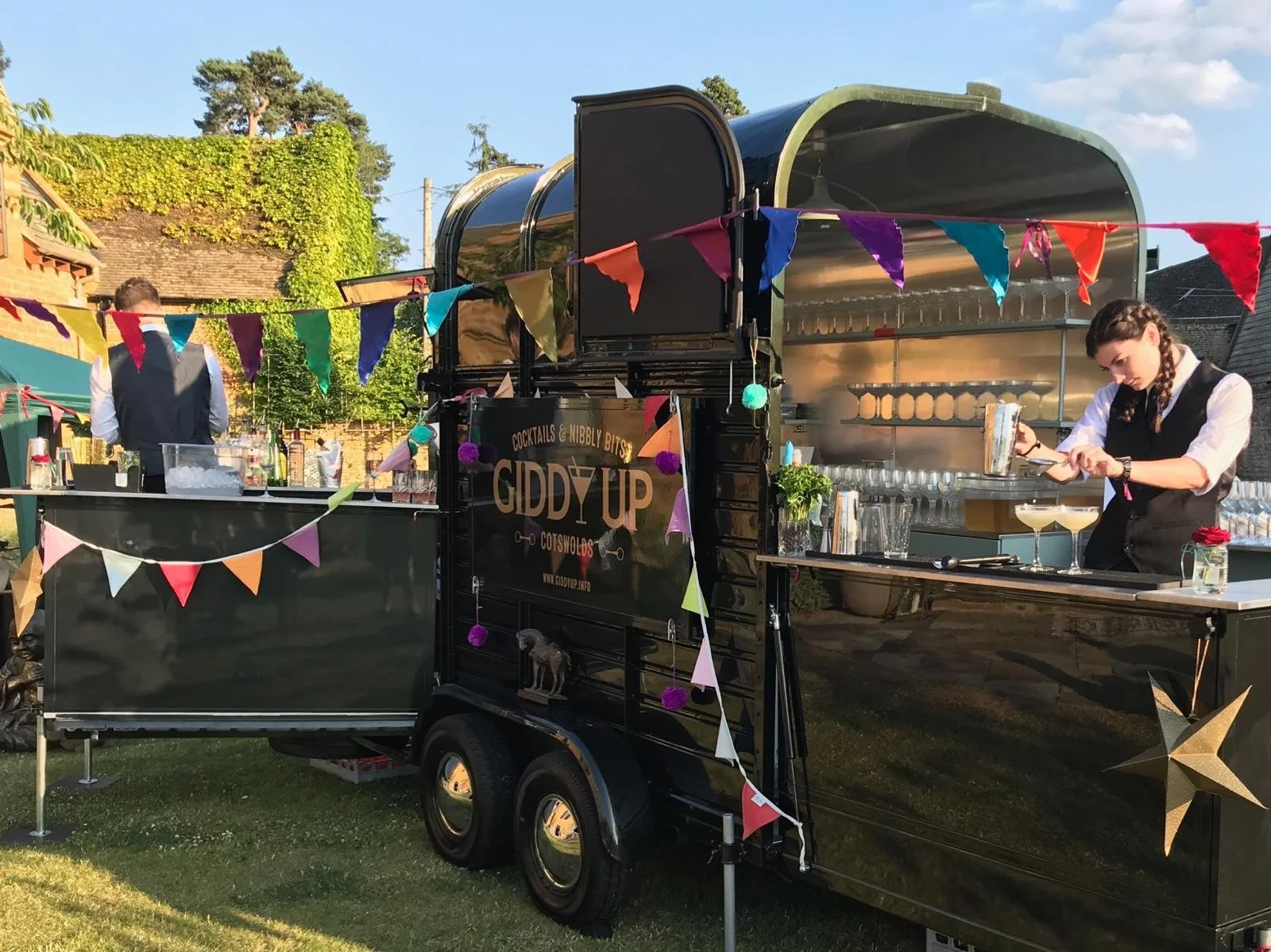 A black mobile bar trailer decorated with colorful bunting and stars, serving drinks at an outdoor event with a woman bartender preparing cocktails and a man standing in the background. Trees and a house with a roof covered in greenery are visible in