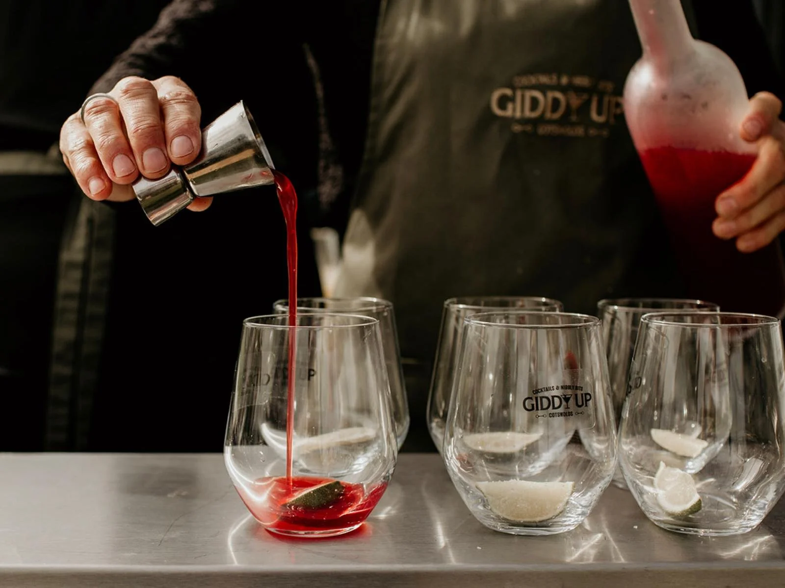 A bartender pouring red liquid into a glass with a lime wedge, while holding another glass with ice and a lime wedge nearby. There are several empty glasses on the counter, with the 'GIDDY UP' logo, some with lime wedges inside.