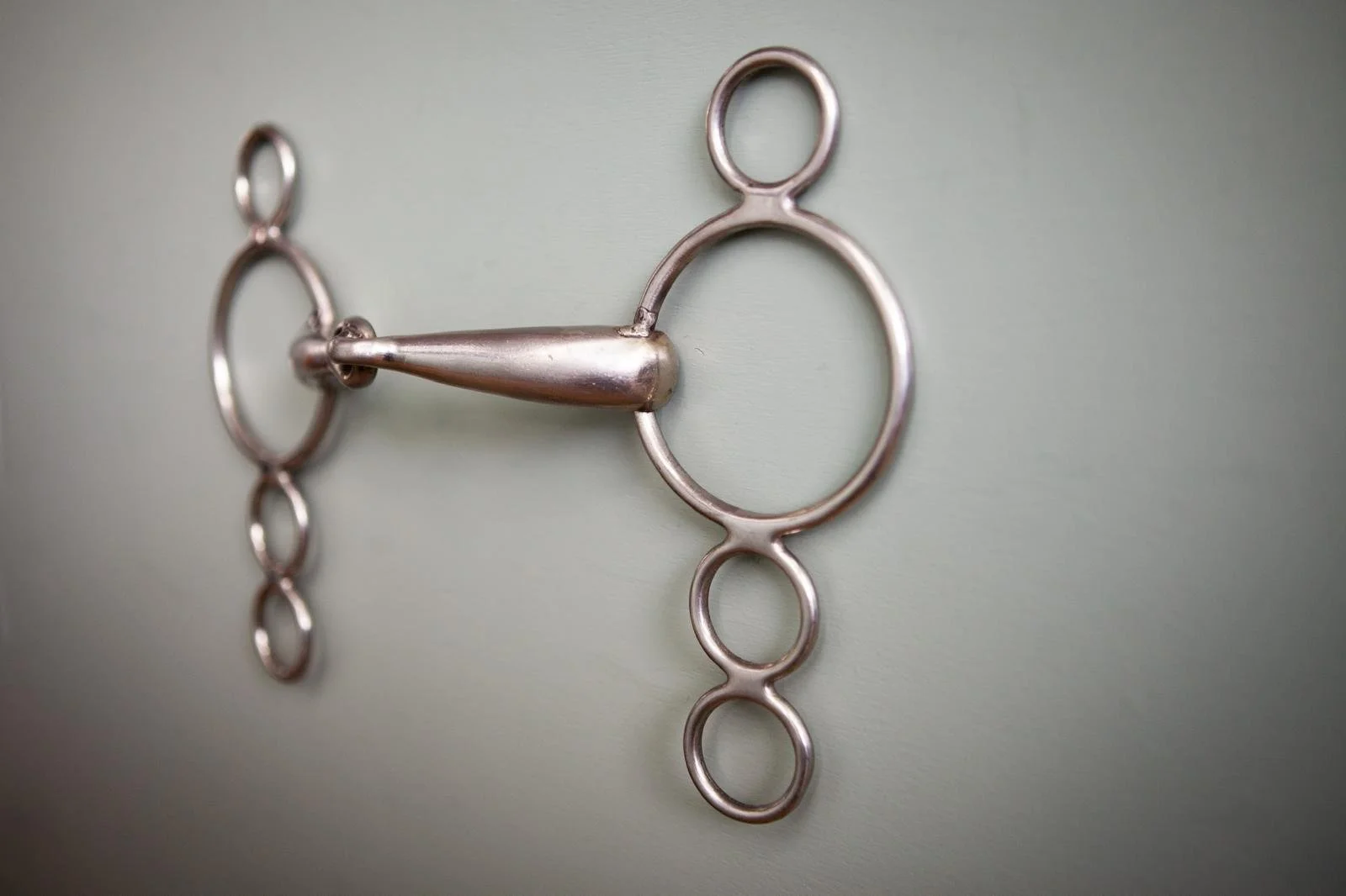 A metallic horse bit with rings on a light green wall, viewed from the side.