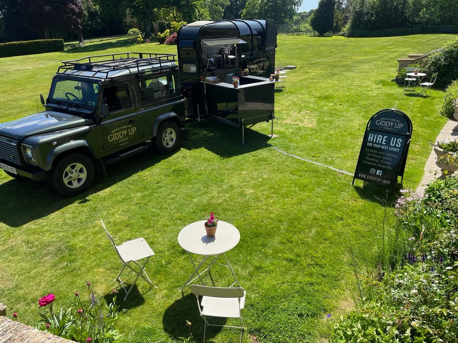 A black catering trailer and a vehicle parked on a lush green lawn, with a sign advertising hiring for events. There are two small white tables with chairs and planters with flowers nearby, with trees and a garden in the background.