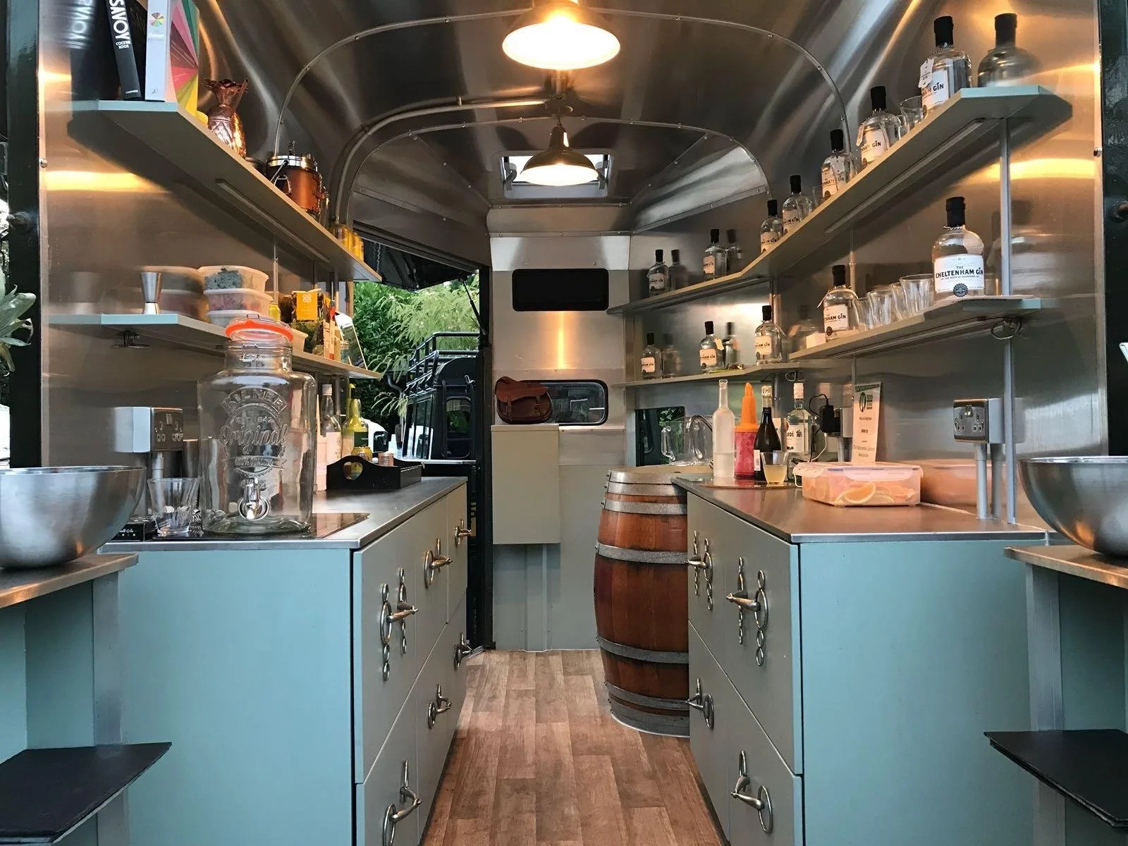 Interior of a mobile bar or food truck with shelves of bottles, bowls, and kitchen supplies, a barrel in the center, and a window with greenery visible outside.