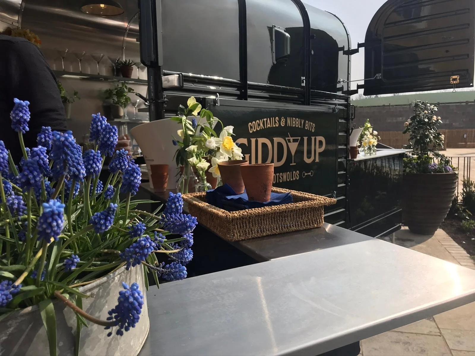 A black mobile bar named Daddy Up with flower decorations on a sunny outdoor patio, with purple and white flowers in pots and a table with a woven basket and small plant pots.