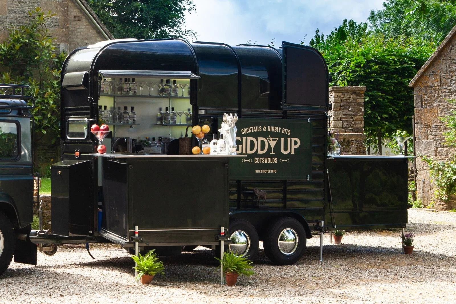 Black mobile bar trailer with the sign Giddy Up, serving cocktails and drinks outdoors, surrounded by potted plants and stone buildings.