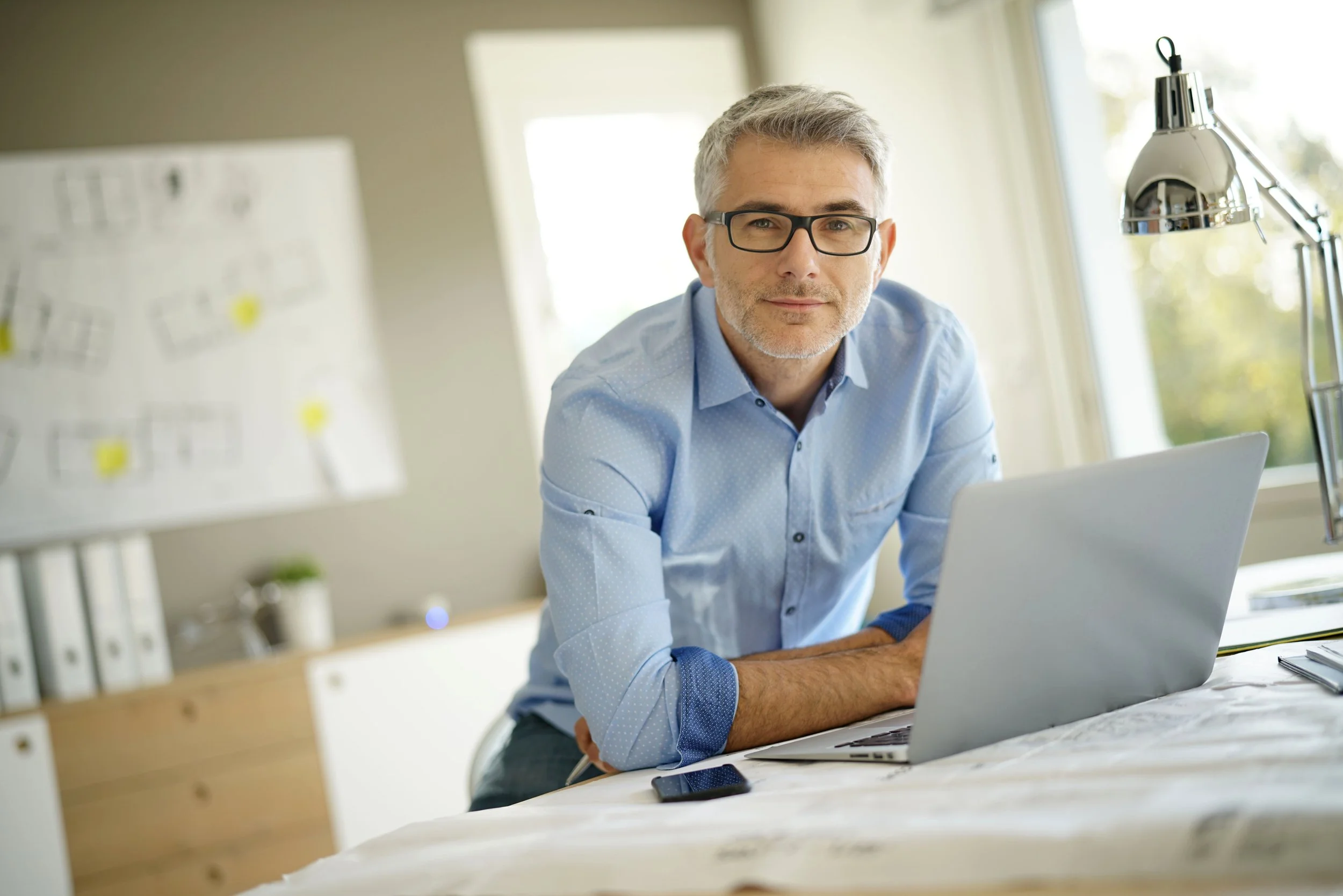 A middle-aged man with gray hair and glasses working on a laptop in a bright office space, with a window in the background and a smartphone on the desk.