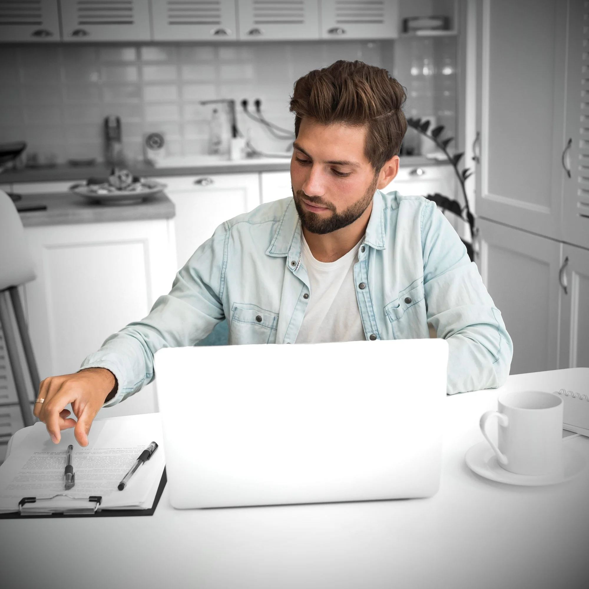 A young man with brown hair and a beard sitting at a kitchen table working on a laptop, with a notebook and pens in front of him, and a mug on his right.