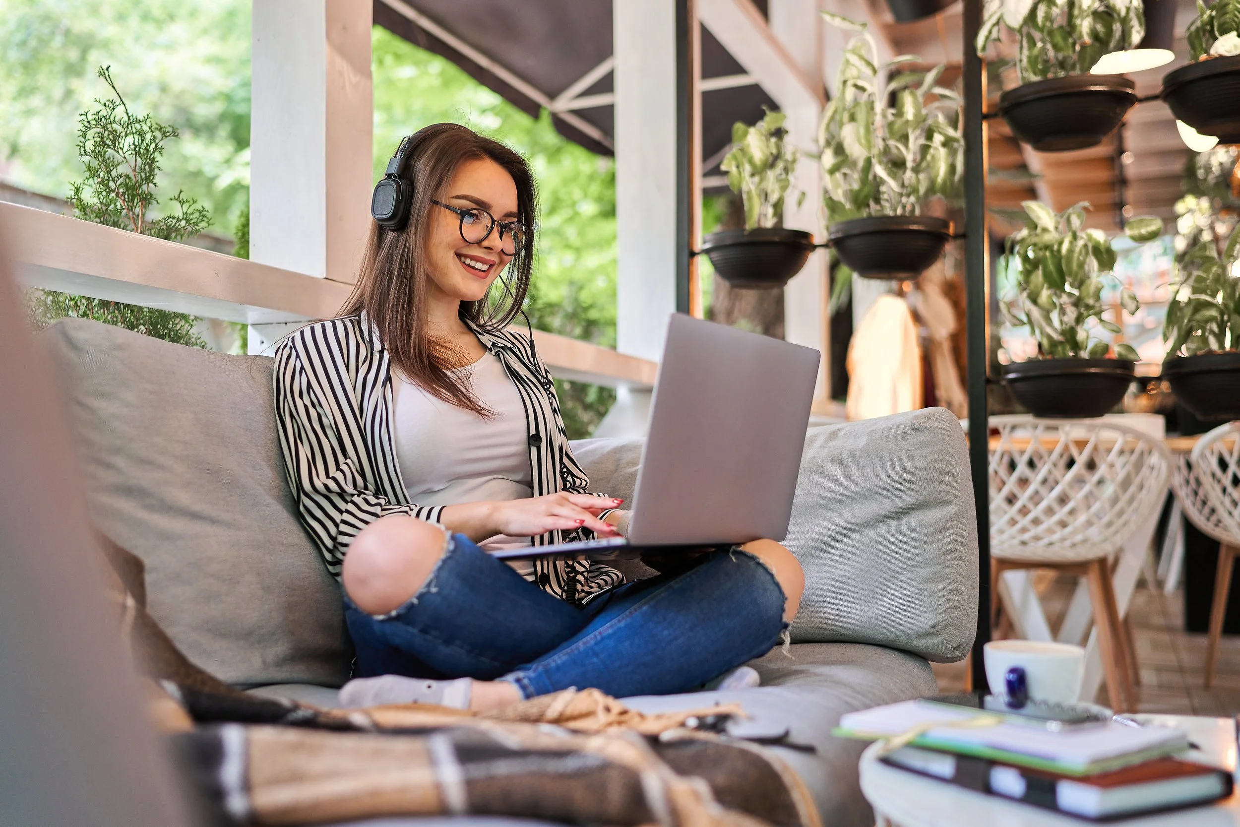 Young woman with headphones sitting on a couch using a laptop, with plants in the background and a table with notebooks and a cup in front of her.