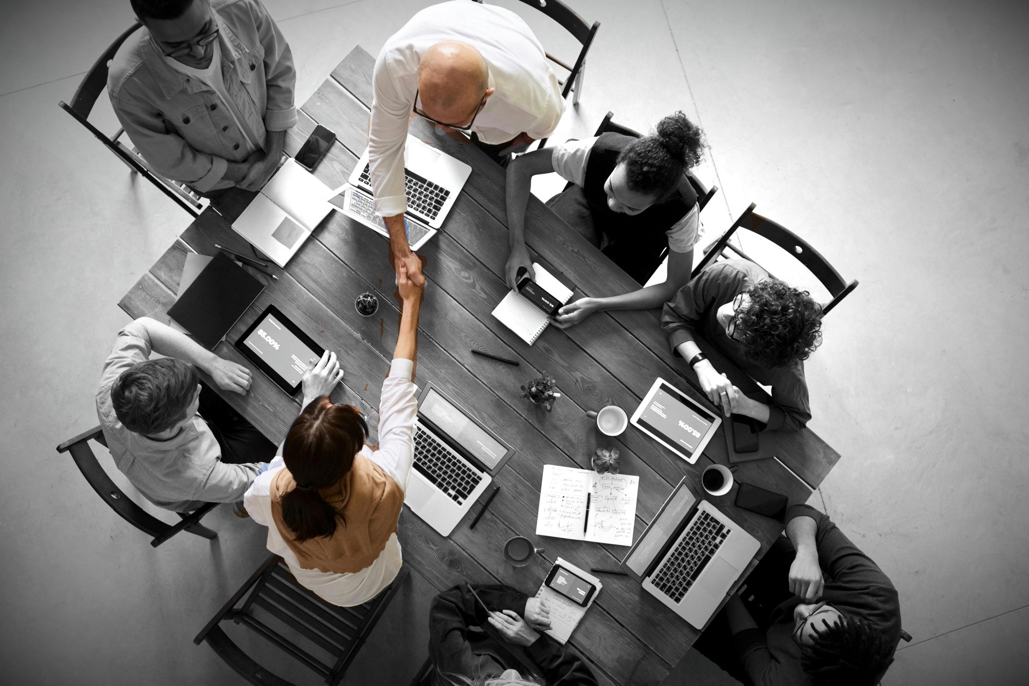 A group of seven people gathered around a wooden table, shaking hands in the center, with laptops, tablets, notebooks, and cups on the table, indicating a business meeting or collaboration.