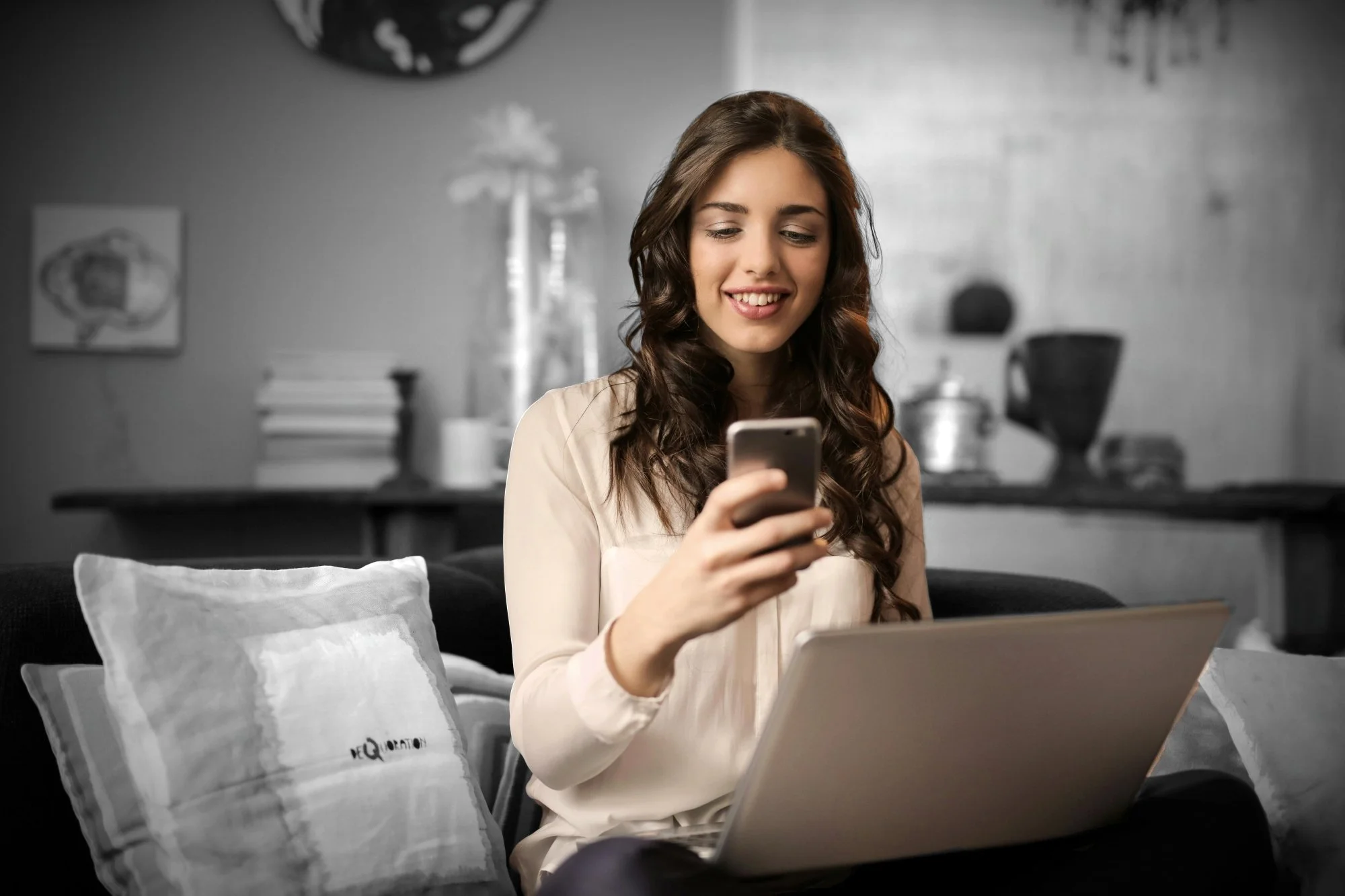 A young woman with long brown curly hair smiling while looking at her smartphone, sitting on a sofa with a laptop on her lap in a cozy living room.