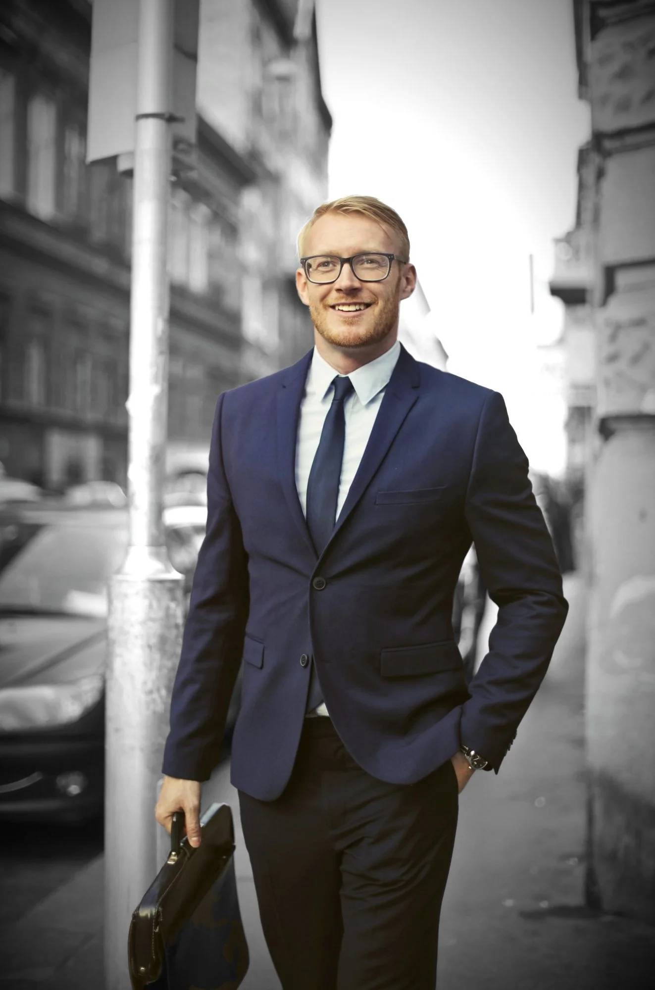 A young man wearing glasses, a dark blue suit, and a tie, holding a briefcase, walking on a city sidewalk with blurred buildings and cars in the background.