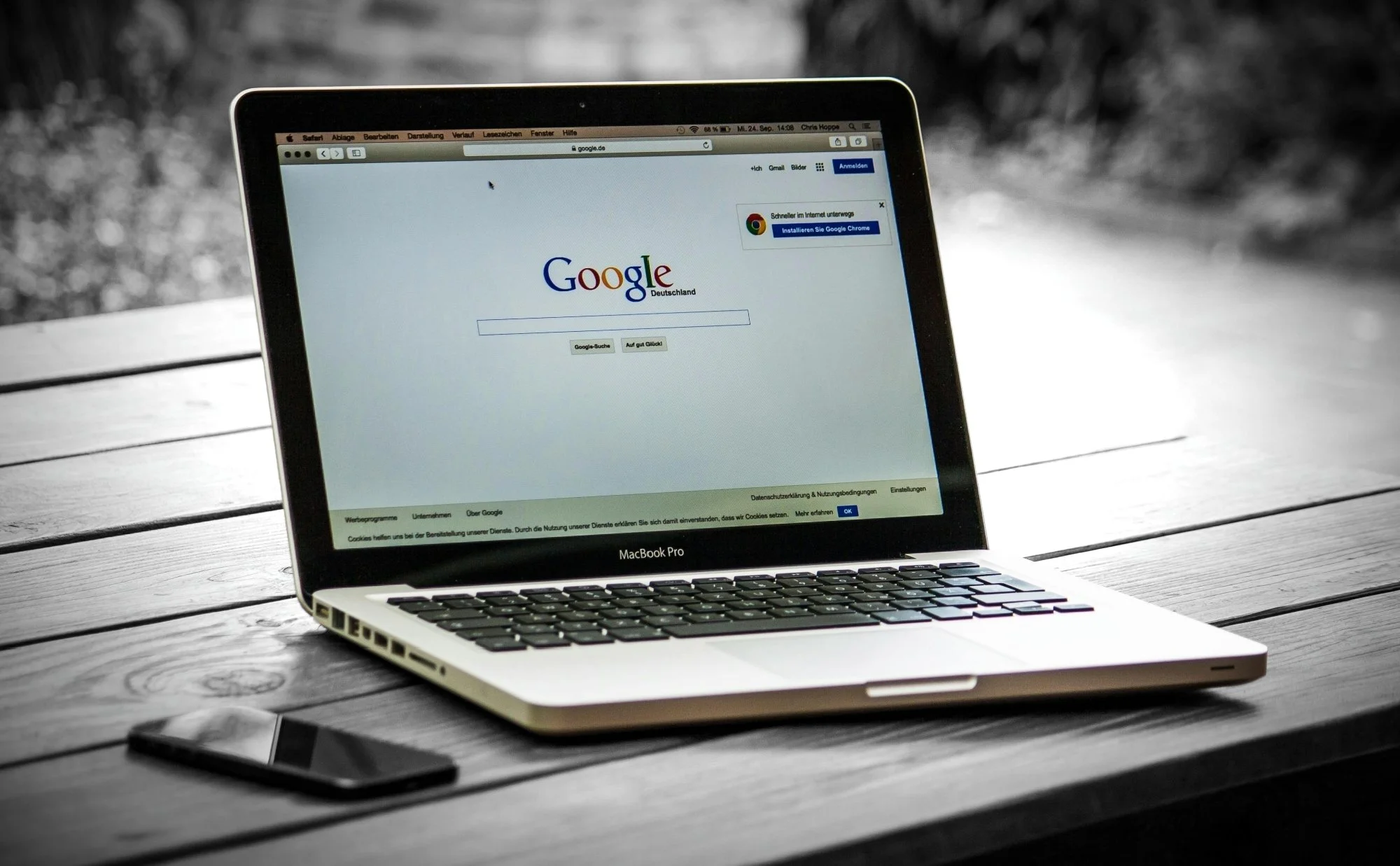 A MacBook Pro laptop on a wooden desk displaying the Google Germany homepage, with a smartphone placed beside it.