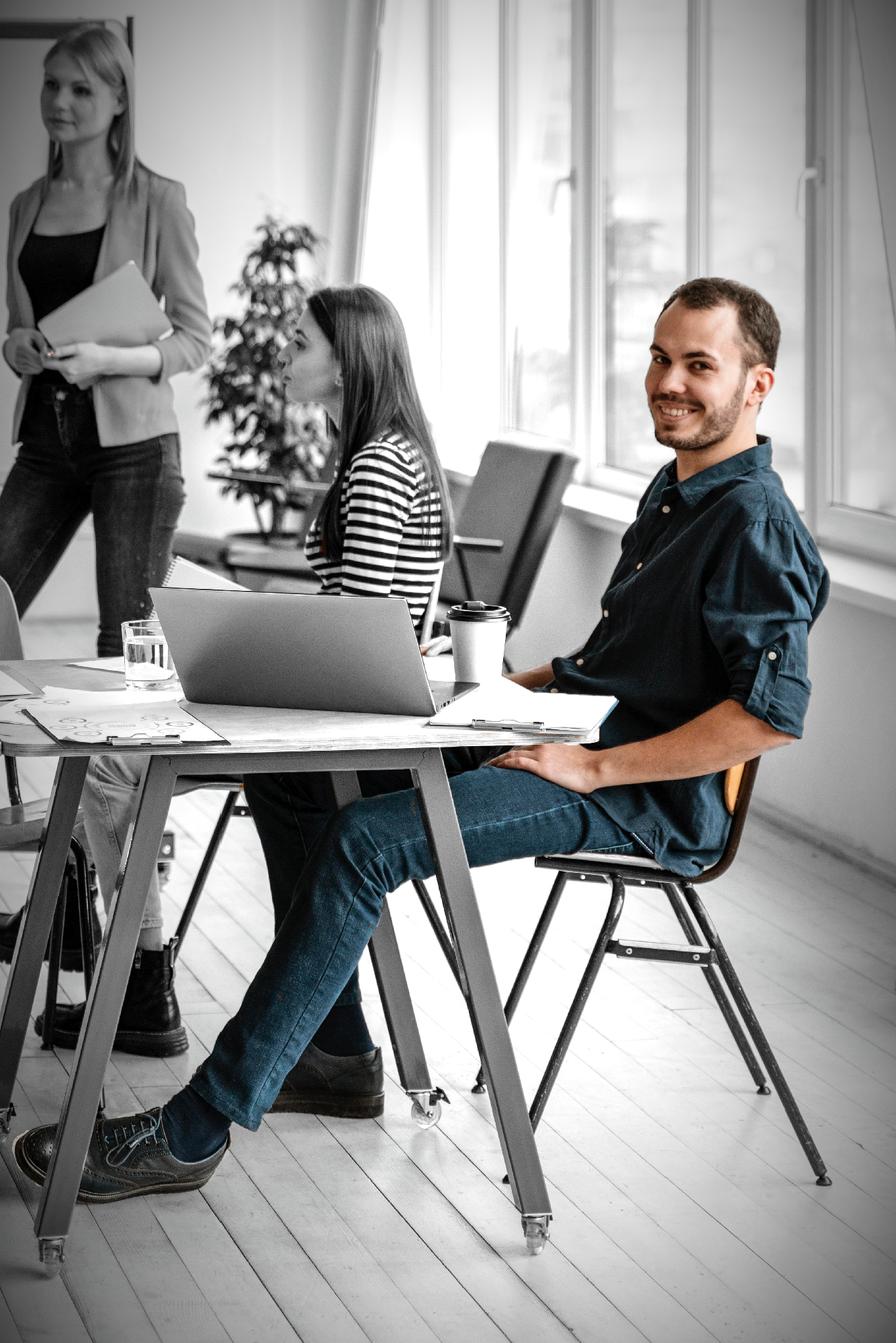 A man smiling and sitting at a table with a laptop and coffee cup, with two women in the background during a meeting in a bright office.