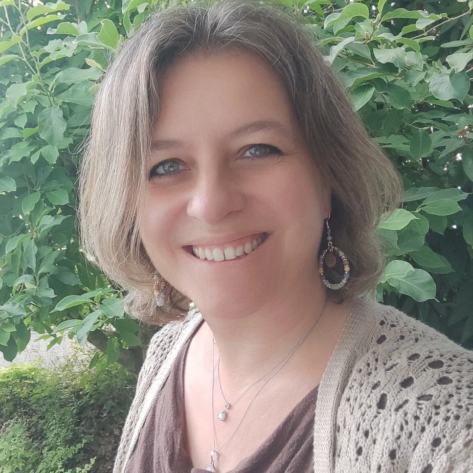 Close-up photo of a woman smiling outdoors, with green leafy plants in the background. She has shoulder-length brown hair, blue eyes, and is wearing earrings, necklaces, a brown top, and a crocheted beige cardigan.
