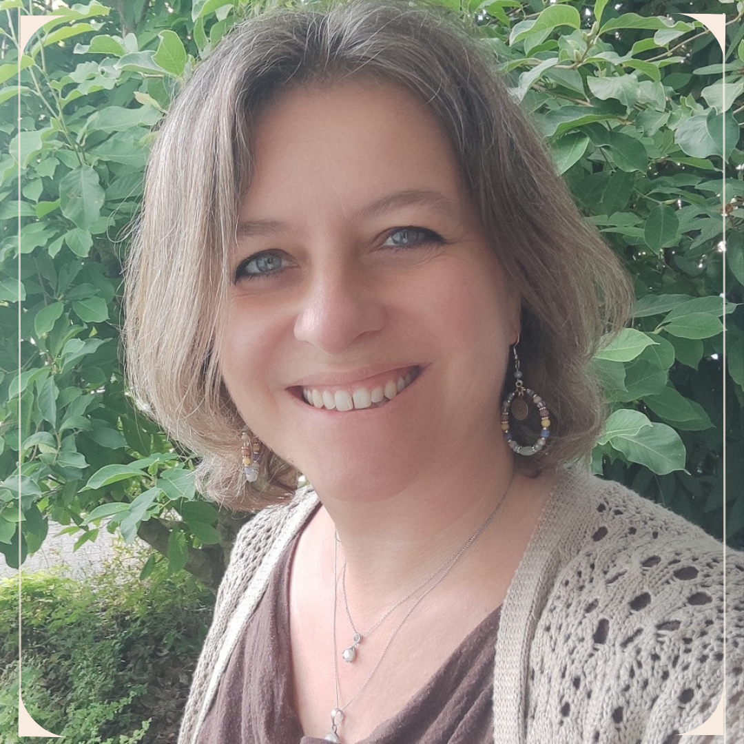 A woman with shoulder-length brown hair, blue eyes, and light skin smiling outdoors in front of green leafy bushes. She is wearing earrings, layered necklaces, and a beige knitted cardigan over a brown top.