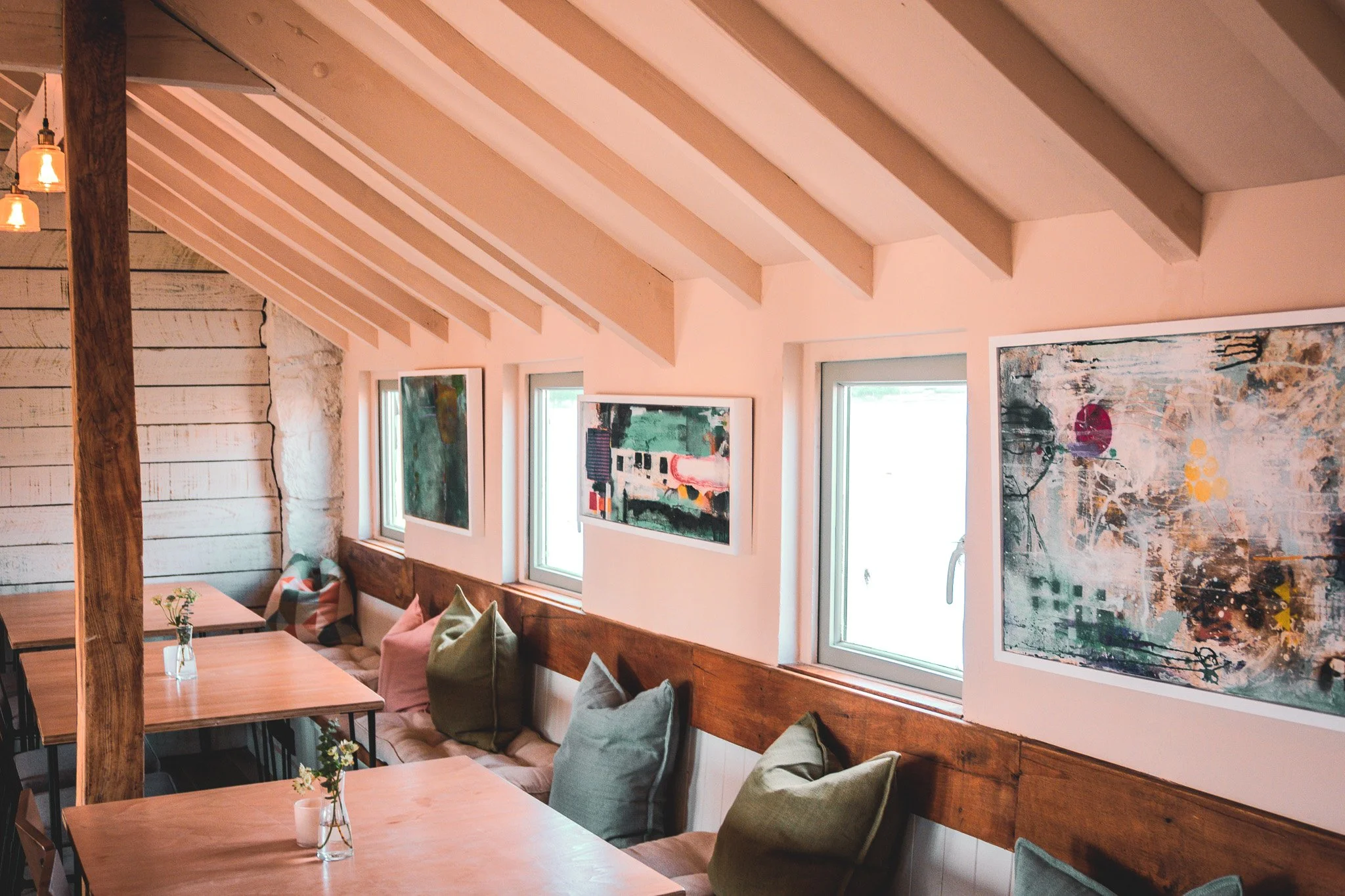 Dinner tables and benches, with comfy cushions and modern artwork, with beams and windows facing the open water