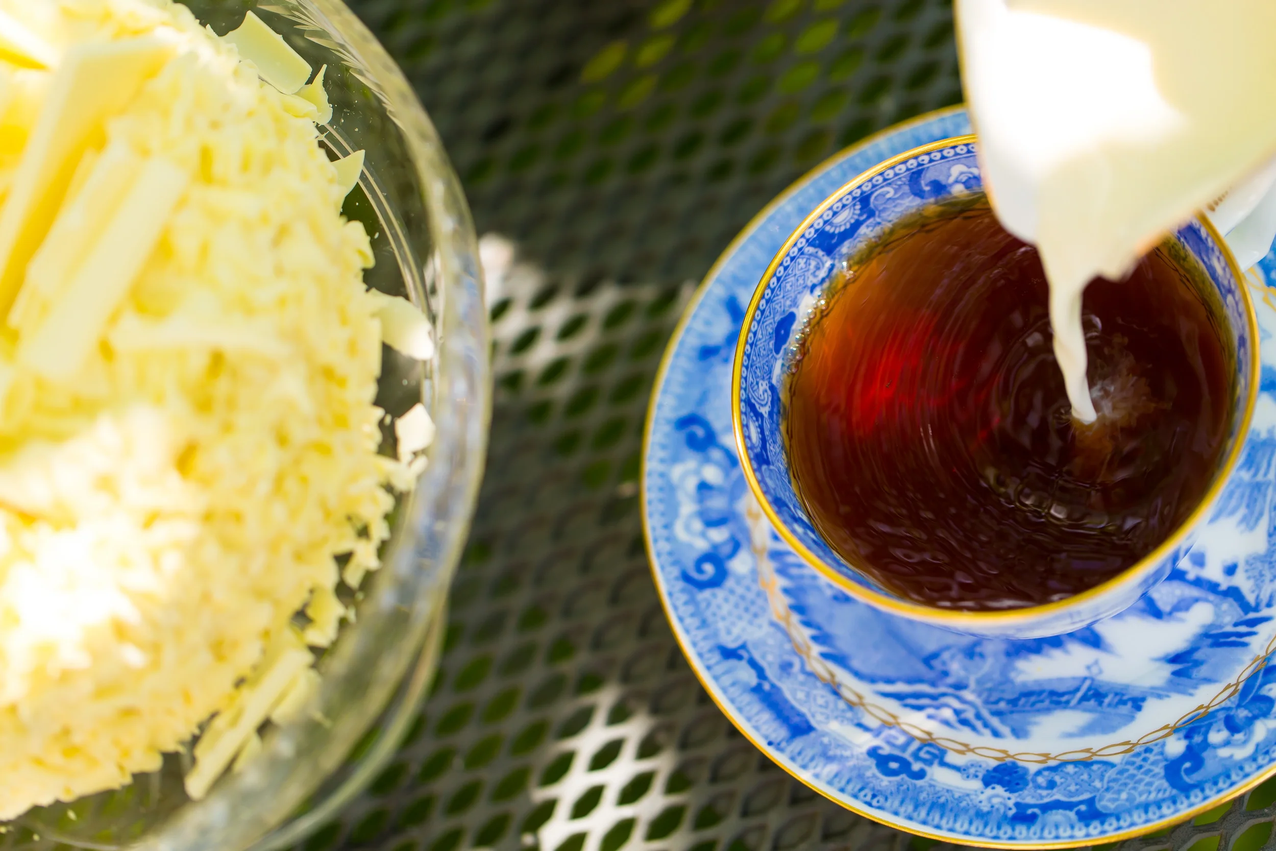 Pouring milk into a cup of tea with a side of white chocolate cake in a glass bowl on a table.