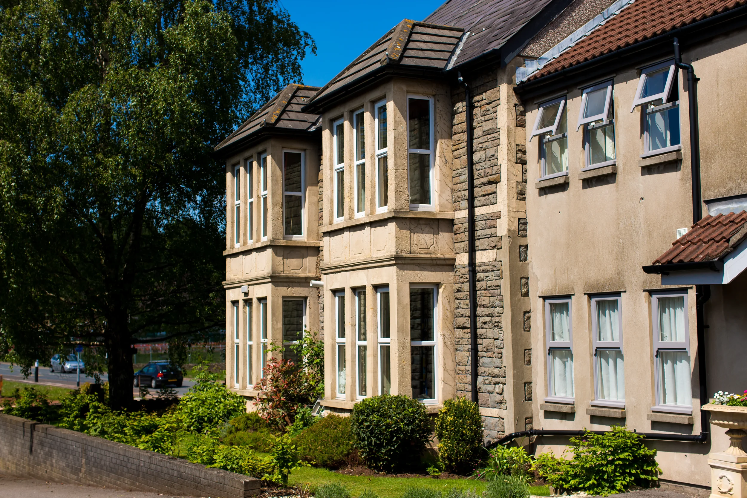 A multi-story residential building with bay windows and a beige stucco exterior surrounded by green bushes and trees.