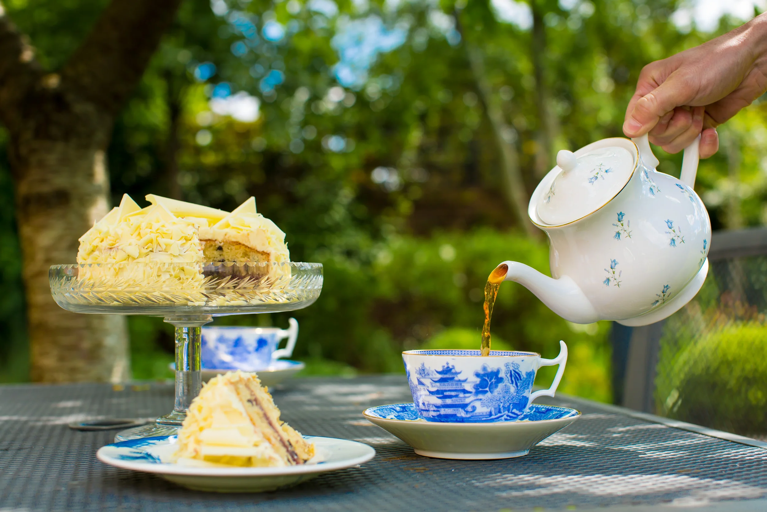 Tea being poured into a cup on an outdoor table with a yellow cake on a stand and a slice on a plate in the foreground, set against a background of green trees.
