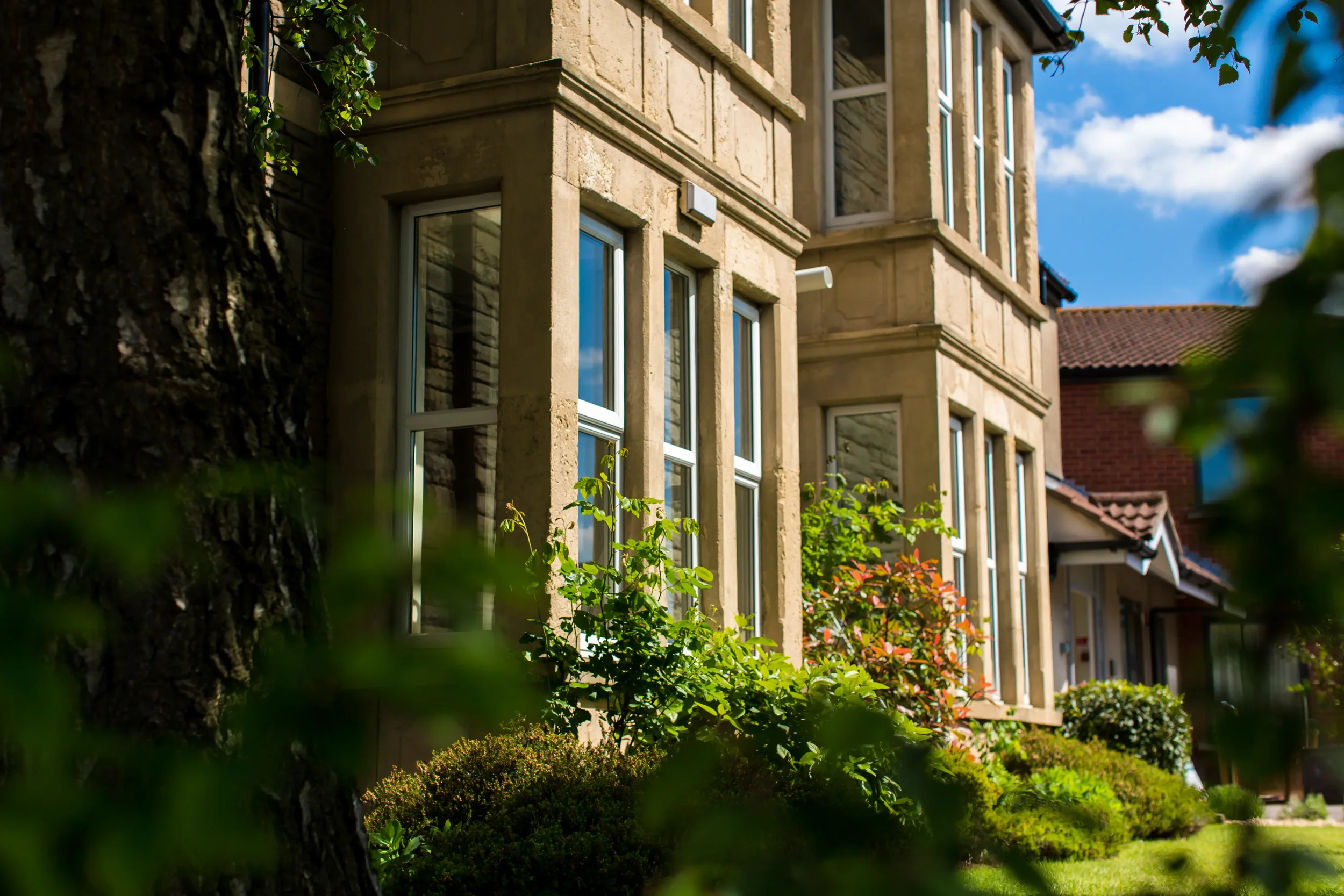 Residential care home building with stone facade, multiple windows, surrounded by greenery, trees, bushes, and a blue sky.